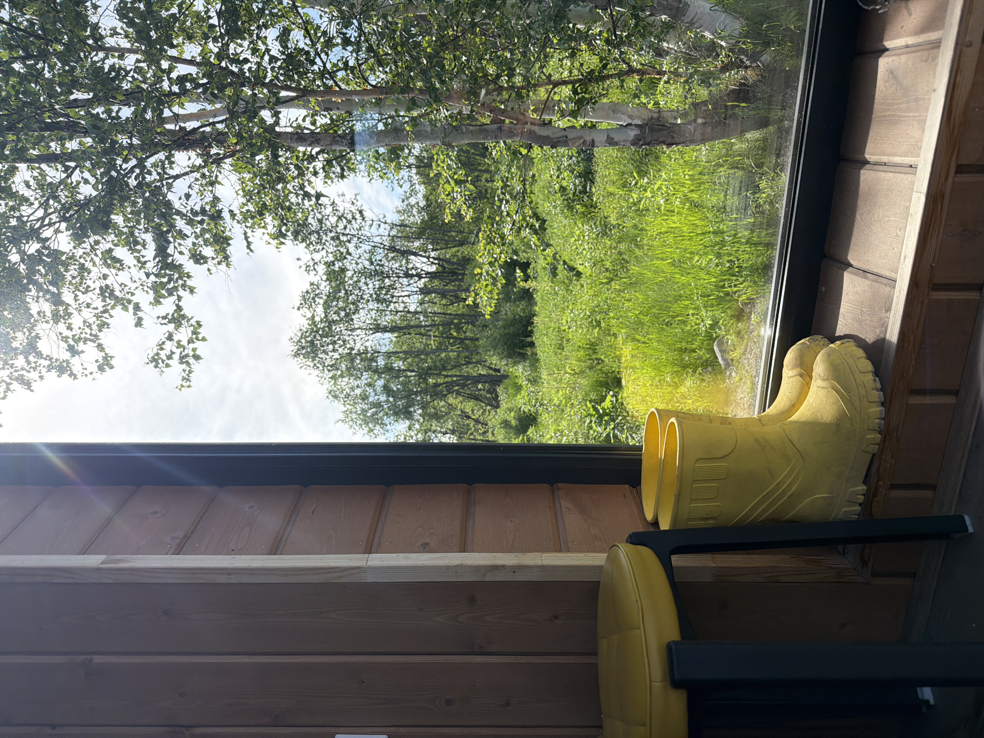The image depicts an interior view from a room with wooden paneling, looking out a large window at a natural outdoor environment. Inside the room, a yellow stool with a tufted seat is partially visible on the left, and a pair of yellow rubber boots rests on a wooden windowsill. The window looks out onto a vibrant, sunlit landscape of tall green grass, numerous trees, and a bright, partly cloudy sky. The scene suggests a rustic or natural setting, possibly a cabin or a rural dwelling, and the presence of the boots implies recent outdoor activity. The time of day appears to be daytime, with strong sunlight casting shadows and creating lens flares in the upper left portion of the image. The location is identified as Novi, Russia.
