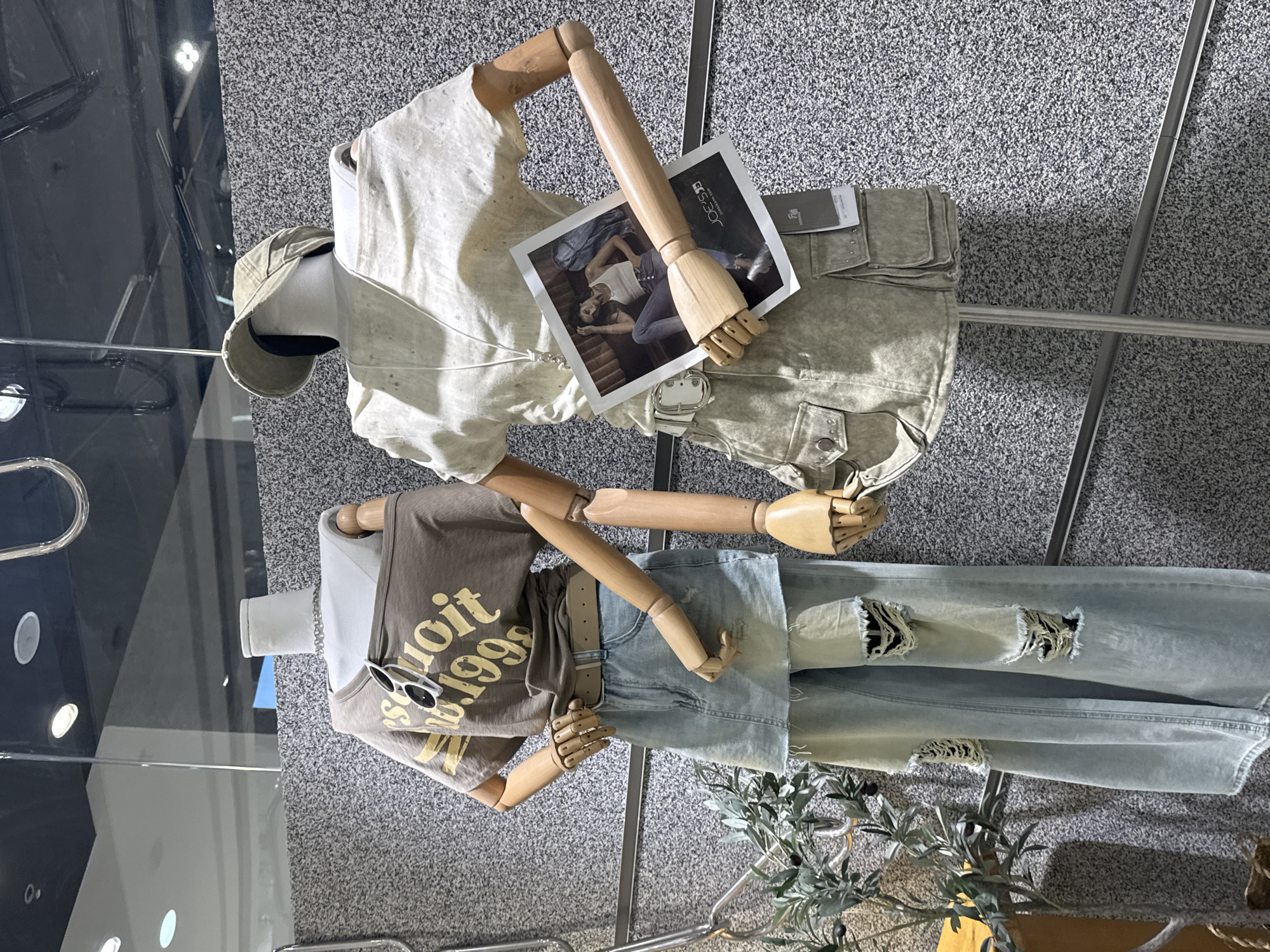 The image shows two mannequins displaying clothing in what appears to be a retail store. The background is a gray, textured surface suggesting a wall or flooring. The top mannequin is wearing a light-colored linen shirt and khaki pants with a belt. It's holding a small magazine or booklet with an image of a person on it. The bottom mannequin is wearing a brown T-shirt with yellow text, light blue denim shorts, and distressed blue jeans. Sunglasses are placed on the shirt. There are metal clothing racks. A plant is to the right of the jeans. There is not enough information to determine if the image is taken in Taihe District, China.