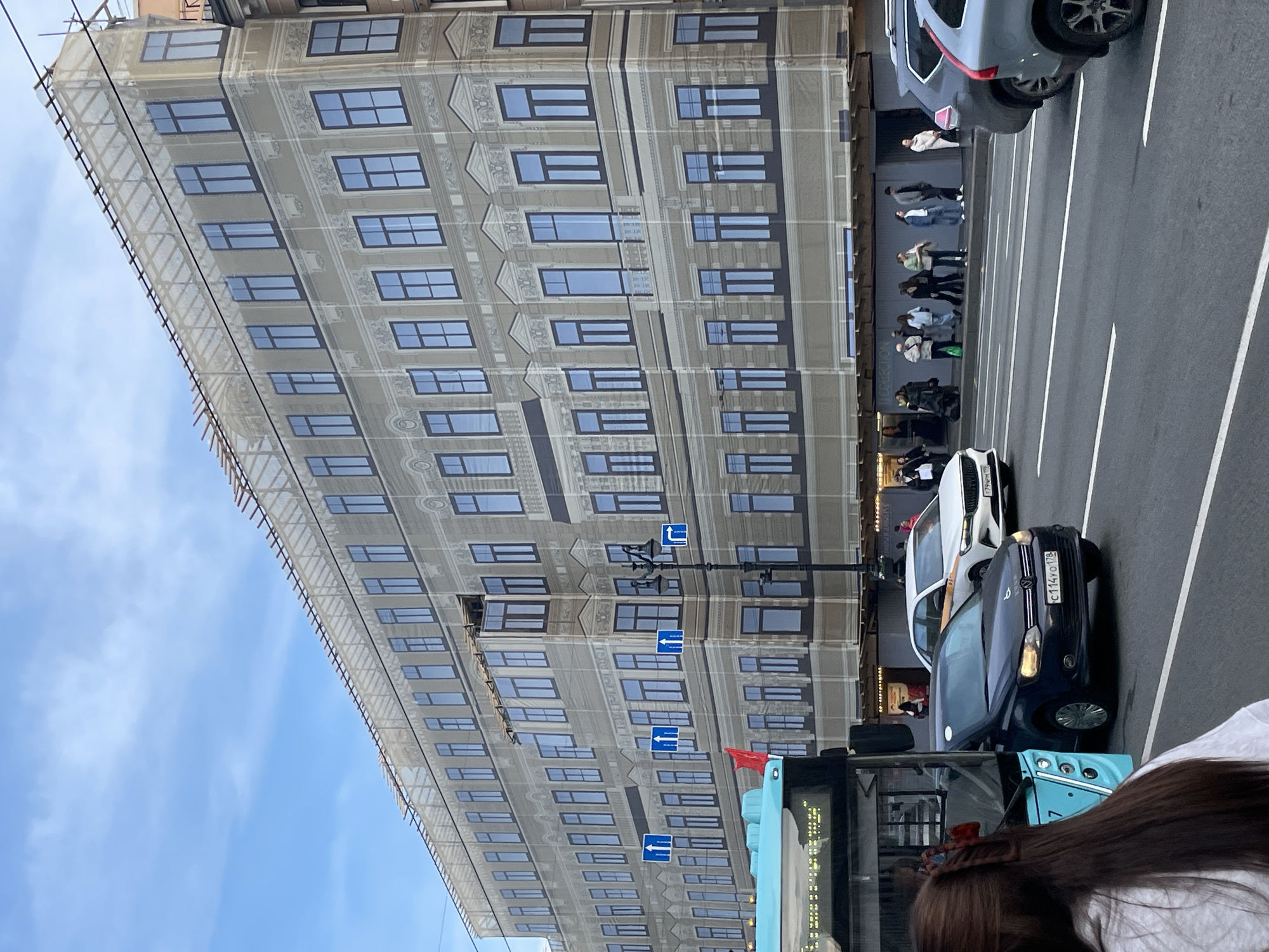 The image depicts a street scene in Saint Petersburg, Russia. A tall building is visible with its facade covered in a mesh or scrim printed with an image of the building itself, potentially indicating ongoing construction or renovation work. The blue sky is visible in the background. A street runs parallel to the building, with a bus and multiple cars traveling on it. Pedestrians are walking along the sidewalk adjacent to the building. Streetlights bearing multiple directional signs are present. A blue city bus is in the foreground, partially obscuring the view of a dark blue car. A woman's hair is visible in the foreground, likely an unintentional inclusion. The license plate on the dark blue car reads 'C114YO 178'. A white SUV is in the background.