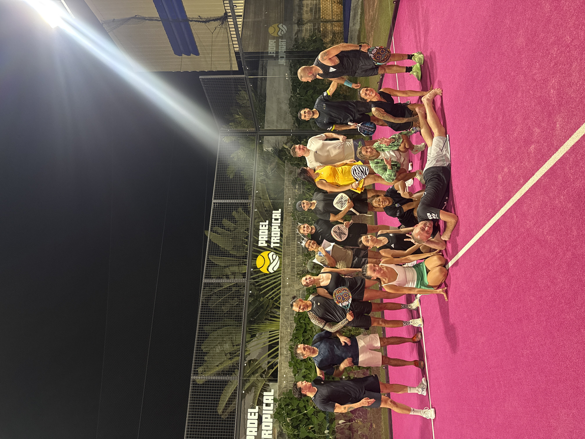 In Ko Samui District, Thailand, a group of eighteen people, consisting of eleven males and seven females, are gathered on a brightly lit pink padel court at night. The group appears to be posing for a photograph, with some standing and others kneeling or lying on the court.
In the standing group, from left to right, there are seven individuals. The first male on the left is wearing a black t-shirt and shorts. Next to him is another male in a navy blue t-shirt and white shorts. Following him is a male in a black and leopard-print long-sleeved shirt and black shorts, holding a padel racket. Beside him stands a female in a black tank top and black shorts. Next, a female is wearing a tan t-shirt and black shorts, holding a diploma. To her right is a male in a dark t-shirt and black shorts, wearing a crown. Finally, the last standing male on the right is wearing a black t-shirt and black shorts, holding a padel racket.
In the front row, from left to right, there are four individuals. A female is kneeling in a white tank top and a green skirt. Next to her, a female is crouching, wearing a green and white checkered shirt over a white t-shirt and white shorts. To her right, a female is on all fours in a black tank top and black shorts. The last person in the front row, a male, is lying on his stomach on the court, wearing a black t-shirt and grey shorts.
Behind the standing group, there are several other individuals partially visible. A male in a black t-shirt and black shorts is standing on the far left. Next to him, a male is wearing a black t-shirt and black shorts. Following him is a male in a white t-shirt and blue patterned shorts, holding a padel racket. To his right, a male in a black t-shirt and black shorts is gesturing with his hand.
The background features a black mesh fence and lush green tropical foliage, suggesting an outdoor setting. The court is marked with white lines. Above the fence, a building structure with blue accents is visible. The lighting is bright, with a strong light source emanating from the upper right of the frame, casting long shadows on the court. The overall atmosphere appears casual and celebratory.