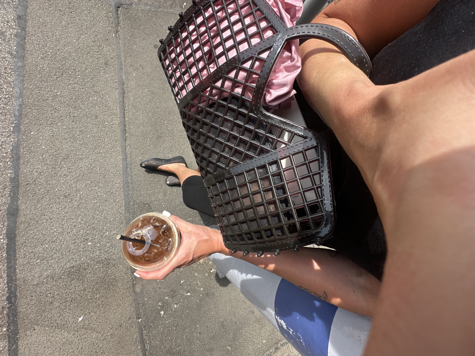 The image, taken from an elevated perspective, depicts a person walking on a street in Tunbridge Wells, United Kingdom. The surface is a mix of concrete and grey asphalt, potentially a sidewalk or pedestrian area adjacent to a road. The person is holding a transparent plastic cup filled with a beverage, likely iced coffee or tea, containing ice and a black straw. They're also carrying a black, cage-like purse with a pink lining over their arm. Only the person's arm, hand holding the cup, a glimpse of the foot wearing a patterned flat shoe, and the purse are visible. A pole painted white with a blue stripe is situated in the lower portion of the image, possibly a street sign or traffic pole. The overall impression is of a candid, street-style shot taken on a sunny day.