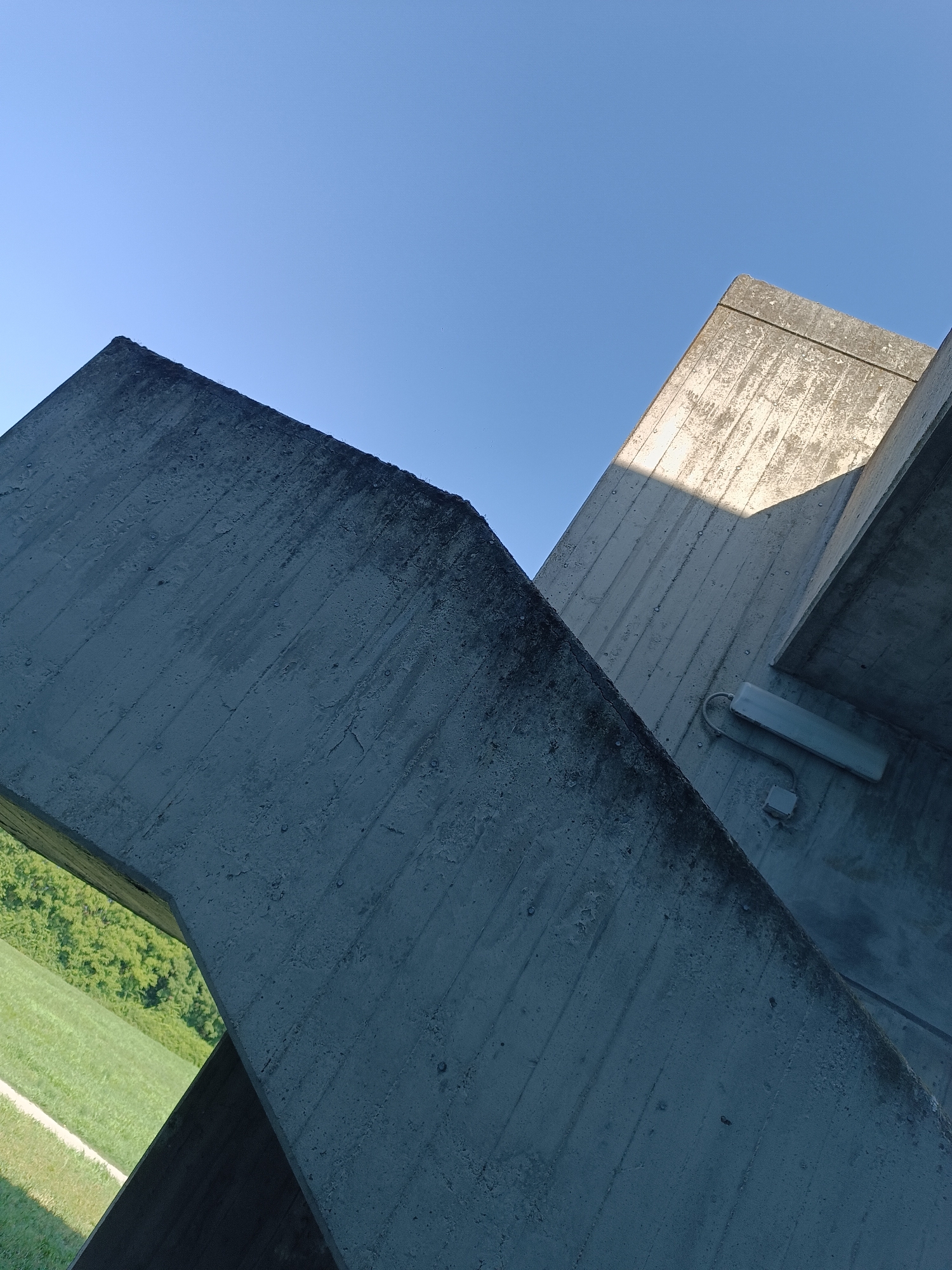 The image displays a low-angle perspective of brutalist concrete structures against a clear blue sky. Two intersecting concrete beams form an 'X' shape, with the foreground beam darker and showing signs of weathering and staining. The background beam is lighter, with visible striations from the concrete molding process, and a section is illuminated by sunlight, casting a shadow. A light-colored rectangular fixture with a coiled wire is attached to the surface of this lighter beam, and below it, a smaller, darker square electrical box is visible. In the lower left portion of the image, a glimpse of a green grassy field with a white path is visible through an opening in the concrete structure. The scene is devoid of people or any overt signs of activity.
The concrete structures are identified as part of the Ponte sul Po (Po River Bridge) in Casalmaggiore, Italy. This bridge is known for its distinctive architectural design featuring large, geometric concrete elements.