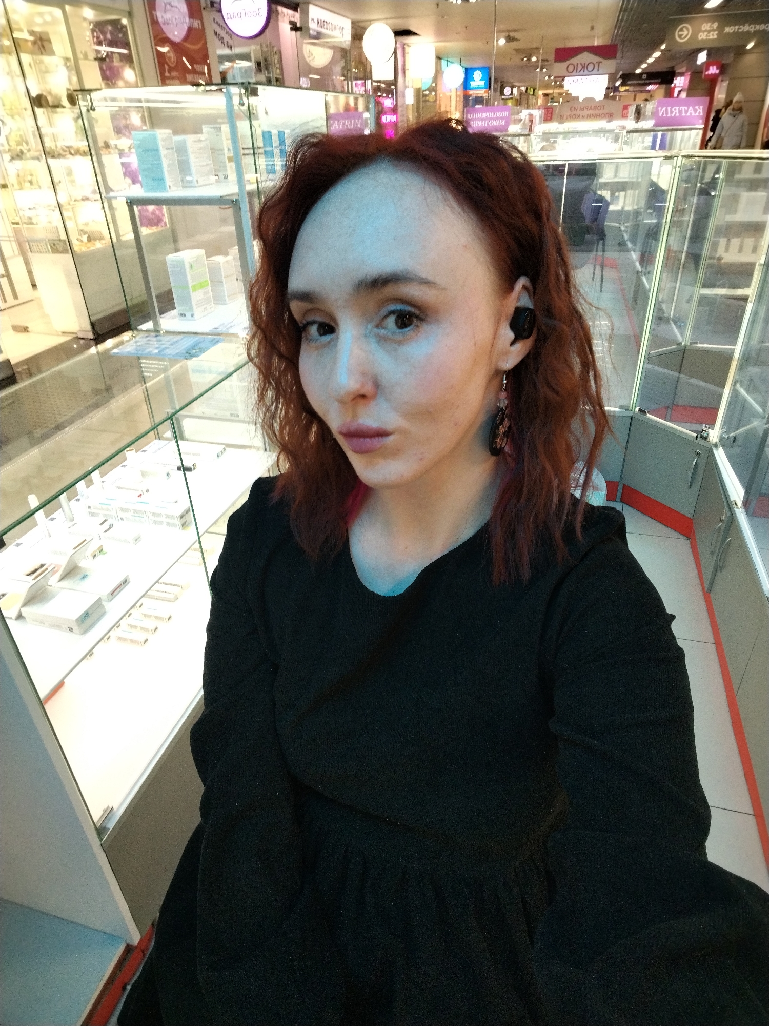 A selfie-style portrait captures a woman with reddish-brown, curly hair and fair skin looking directly at the camera with a neutral to slightly pouting expression. She is wearing a black long-sleeved garment, possibly a dress or top. A black wireless earbud is visible in her right ear, along with a dark, dangling earring.
The setting appears to be an indoor shopping mall or department store in Izhevsk, Russia. The background is filled with glass display cases, showcasing a variety of packaged products, which look like cosmetics, skincare items, or small electronics. Bright artificial lighting illuminates the scene. Reflections of overhead lights and other elements of the commercial environment are visible on the glass.
Notable details in the background include several signs and banners. Clearly visible text includes "TOKYO" (both directly and mirrored) and parts of the Russian name "КАТРИН" (Katrin). Other fragmented or mirrored Russian text, such as "МОЙ ДЕЛА" (My Deeds), can also be discerned on various signs. The floor is tiled, featuring white sections with red or orange accent strips. The overall impression is that of a casual outing in a bustling commercial area.