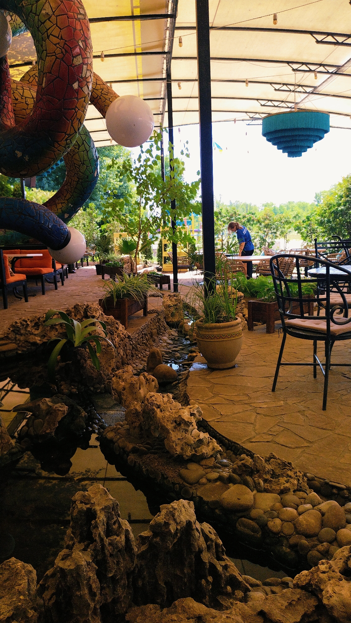 The image depicts an outdoor seating area of a restaurant or cafe, likely in the city of Horishni Plavni, Ukraine, given the context. In the foreground, a man-made stream or pond is lined with large rocks and smaller pebbles, with some greenery in planters. A large, colorful, mosaic-covered serpent sculpture is visible on the left, coiled around a support structure. In the background, a woman in a blue shirt and dark pants is seen standing at a table. The seating area features stone paving, outdoor furniture including tables and chairs, and a covered roof structure. Trees and foliage surround the area, suggesting a park-like or garden setting. A unique, tiered, blue chandelier-like fixture hangs from the ceiling.