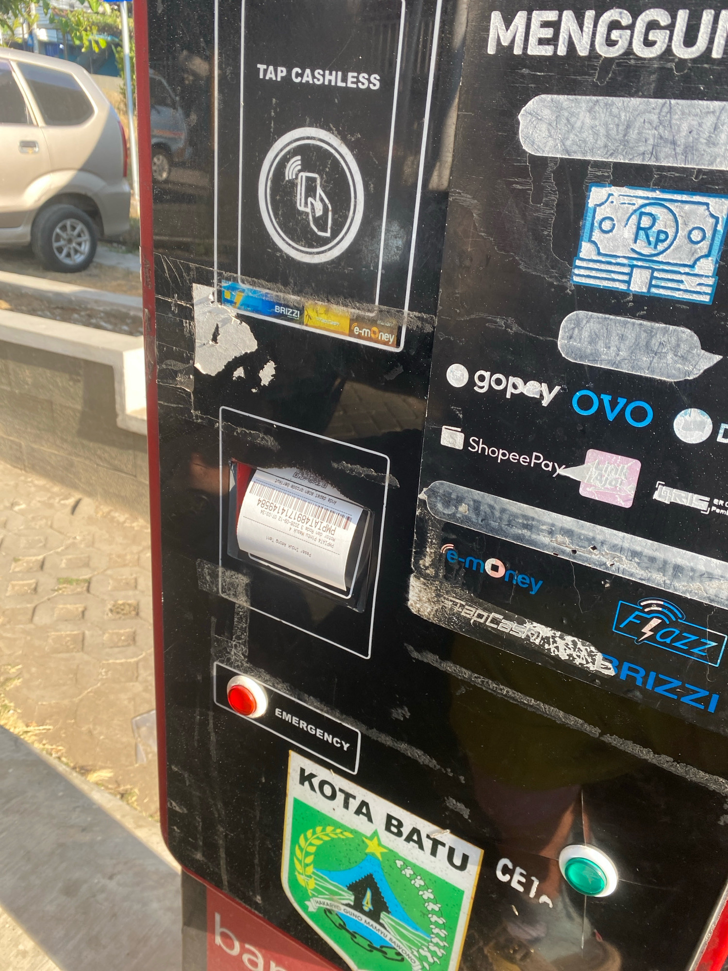 A close-up, eye-level, outdoor, medium shot captures a payment machine in the city of Unknown, Unknown. The machine is black and appears weathered, with numerous scratches and peeling stickers. In the upper left, a silver car is partially visible. The machine displays various payment logos, including "BRIZZI," "e-money," "Gopay," "OVO," and "ShopeePay." A receipt is partially dispensed from a slot on the machine. An "EMERGENCY" button, which is red, is visible on the lower left side of the machine. A city crest for "KOTA BATU" is affixed to the machine on the lower right.