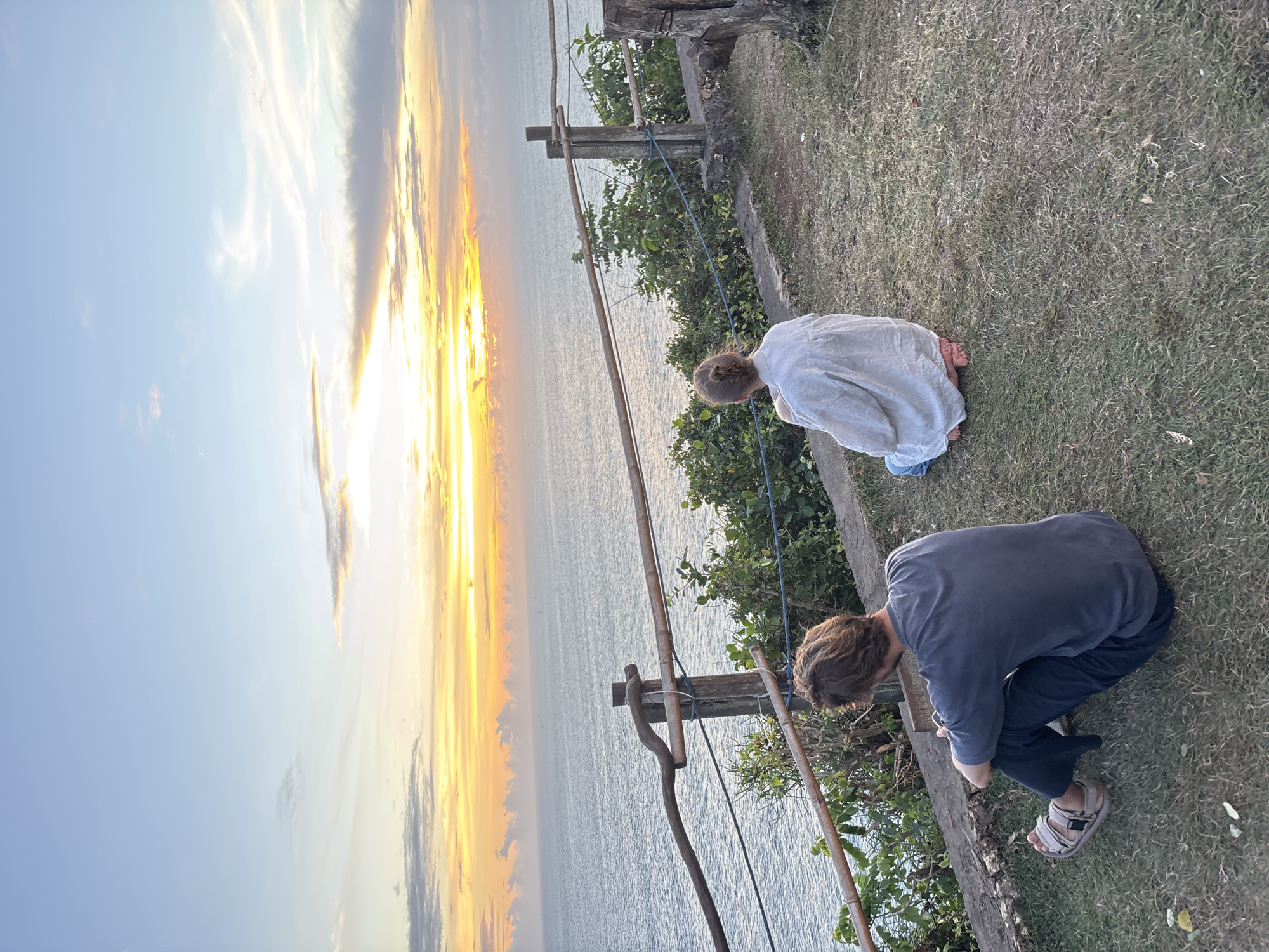 Two people are kneeling at the edge of a grassy area overlooking a body of water and a sunset. The sky is light blue and orange, with a yellow glow. The water is calm. A crude wooden fence is present. The scene appears to be located on a cliff overlooking the ocean. This is likely near Kuta Selatan, Indonesia, due to the landscape and the sunset. The people appear to be observing something below at the edge of the cliff.