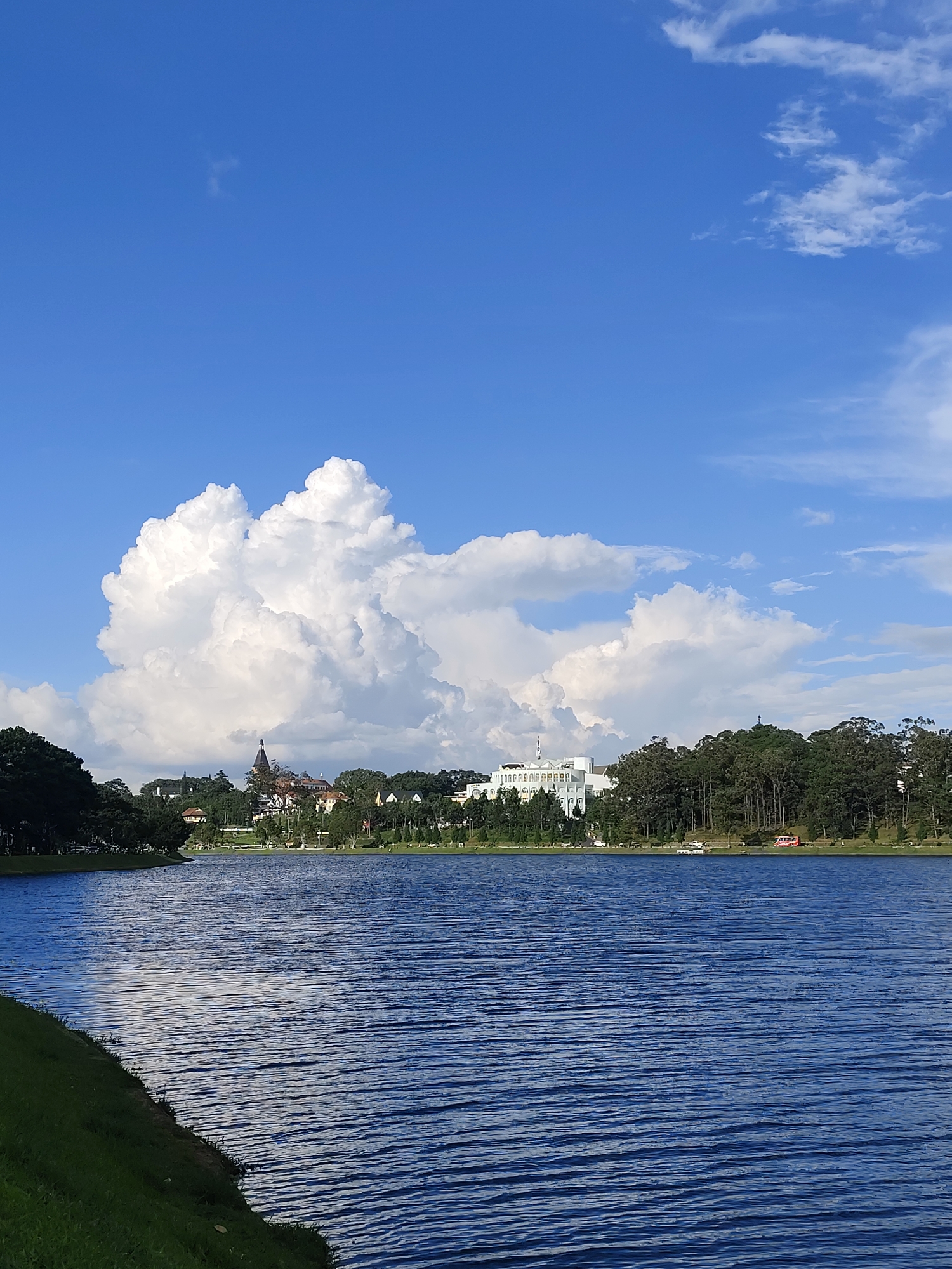 A wide shot captures Xuan Huong Lake in Da Lat, Vietnam, under a bright blue sky dotted with large, fluffy white clouds. The lake's surface is a deep blue, rippling with gentle waves that reflect the light. On the far bank, a line of lush green trees forms a natural border. Behind the trees, several buildings are visible, including a prominent white structure with arches and a building with a dark, pointed steeple. The foreground shows a grassy embankment sloping down to the water's edge. The overall scene conveys a serene and picturesque view of the lake and its surrounding landscape.