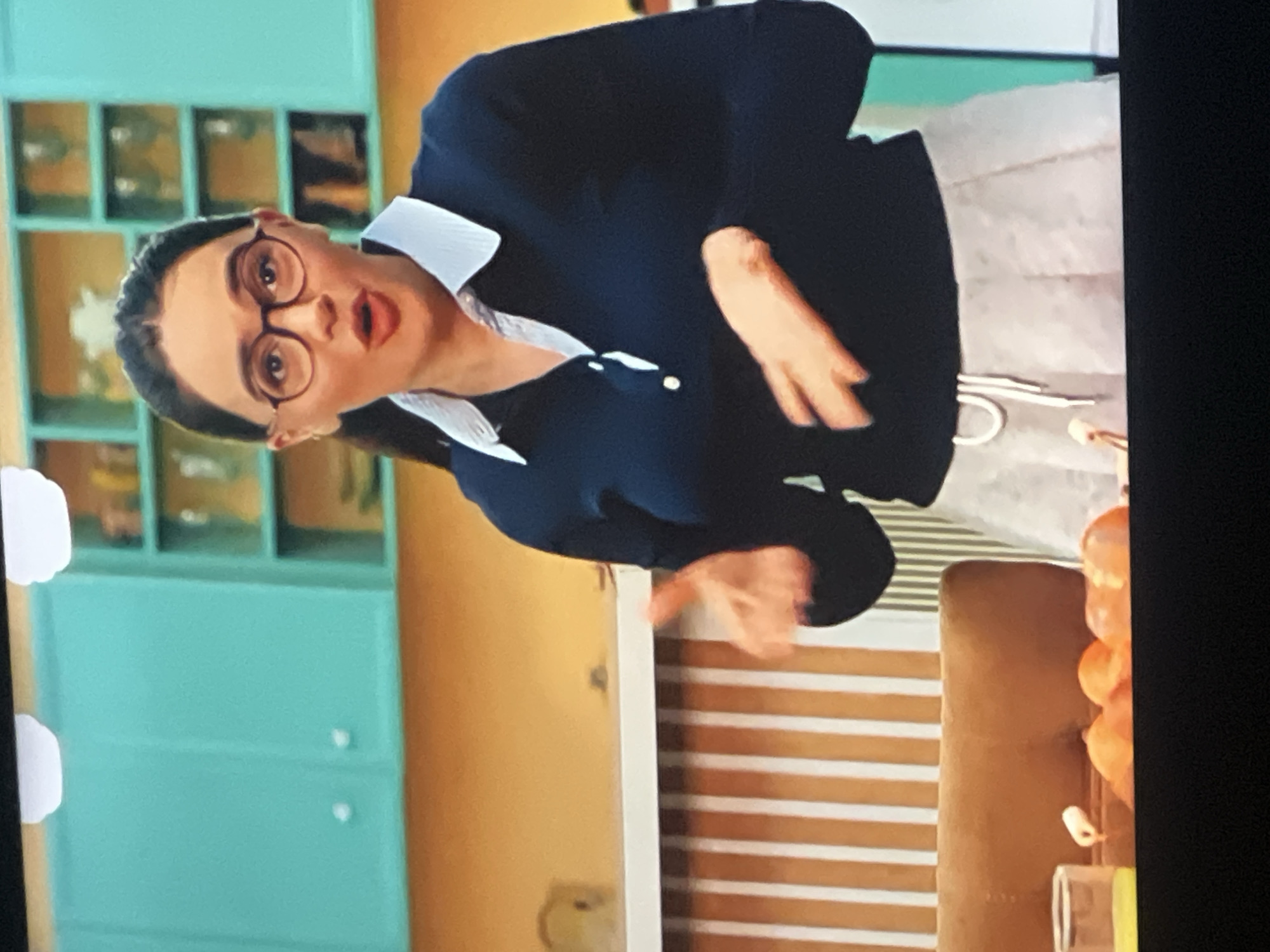 The image shows a woman with dark hair, wearing glasses, a dark blue collared shirt, and a light grey pleated skirt. She is standing in a kitchen with teal cabinets and an orange wall. She is gesturing with her hands, and there are some orange fruits and a glass of yellow liquid on the counter in front of her. The image does not provide any information about a location in the city of Vanadzor, Armenia.
