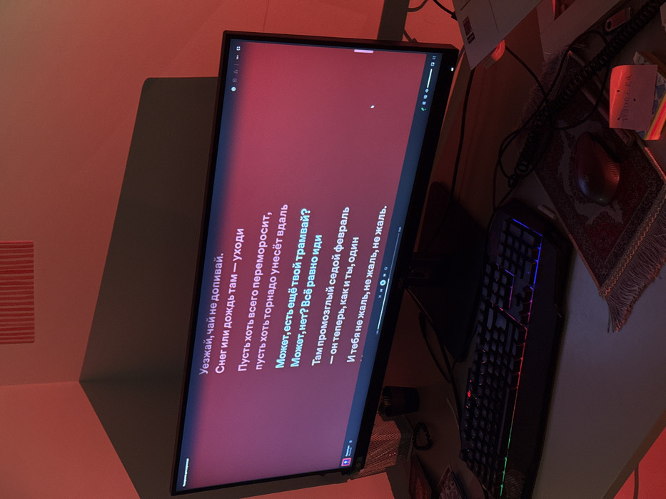 The image captures a desk setup in what appears to be a room with warm, reddish lighting. The scene is illuminated primarily by this ambient light, giving everything a red-orange hue. The main focal point is a computer monitor displaying text against a dark red background.
The monitor is positioned centrally, slightly angled, showing what looks like lyrics or a poem in Russian. The text is white and is presented in several lines, implying a sequence, possibly for karaoke or a text display from a video. The visible text reads:
"Уезжай, чай не допивай.
Снег или дождь там — уходи
Пусть хоть всего переморосит,
пусть хоть торнадо унесёт вдаль
Может, есть ещё твой трамвай?
Может, нет? Всё равно иди
Там промозглый седой февраль
- он теперь, как и ты, один
И тебя не жаль, не жаль, не жаль."
Translated to English, this means:
"Leave, don't finish your tea.
Snow or rain there — just go
Let everything be frozen over,
let a tornado carry you far away
Maybe your tram is still there?
Maybe not? Go anyway
There's a damp, gray February
- it is now, like you, alone
And I don't feel sorry for you, don't feel sorry, don't feel sorry."
At the bottom of the screen, there are media player controls (play/pause, skip, volume) and a progress bar, suggesting that the text is synchronized with an audio or video track. The upper part of the screen shows what appear to be application icons or tabs in a browser.
To the right of the monitor, on the desk, there is a gaming keyboard with multicolored backlighting, predominantly blue, purple, and red. Further to the right, a computer mouse rests on an ornate, patterned mousepad, which has fringes along its edges and a design reminiscent of a small prayer rug. Other items on the desk include some cables, a small piece of paper with handwriting that is difficult to decipher, and possibly a laptop or other electronics in the darker background.
The room's wall to the left of the monitor is painted in a light color, but heavily saturated with the red light, creating a strong orange-red reflection. There's also a subtle striped pattern visible on this wall in the upper left corner, possibly from a textured wallpaper or decor. The overall mood is intimate and somewhat melancholic, amplified by the warm, dim lighting and the nature of the displayed text. The time of day is likely evening or night due to the artificial lighting.