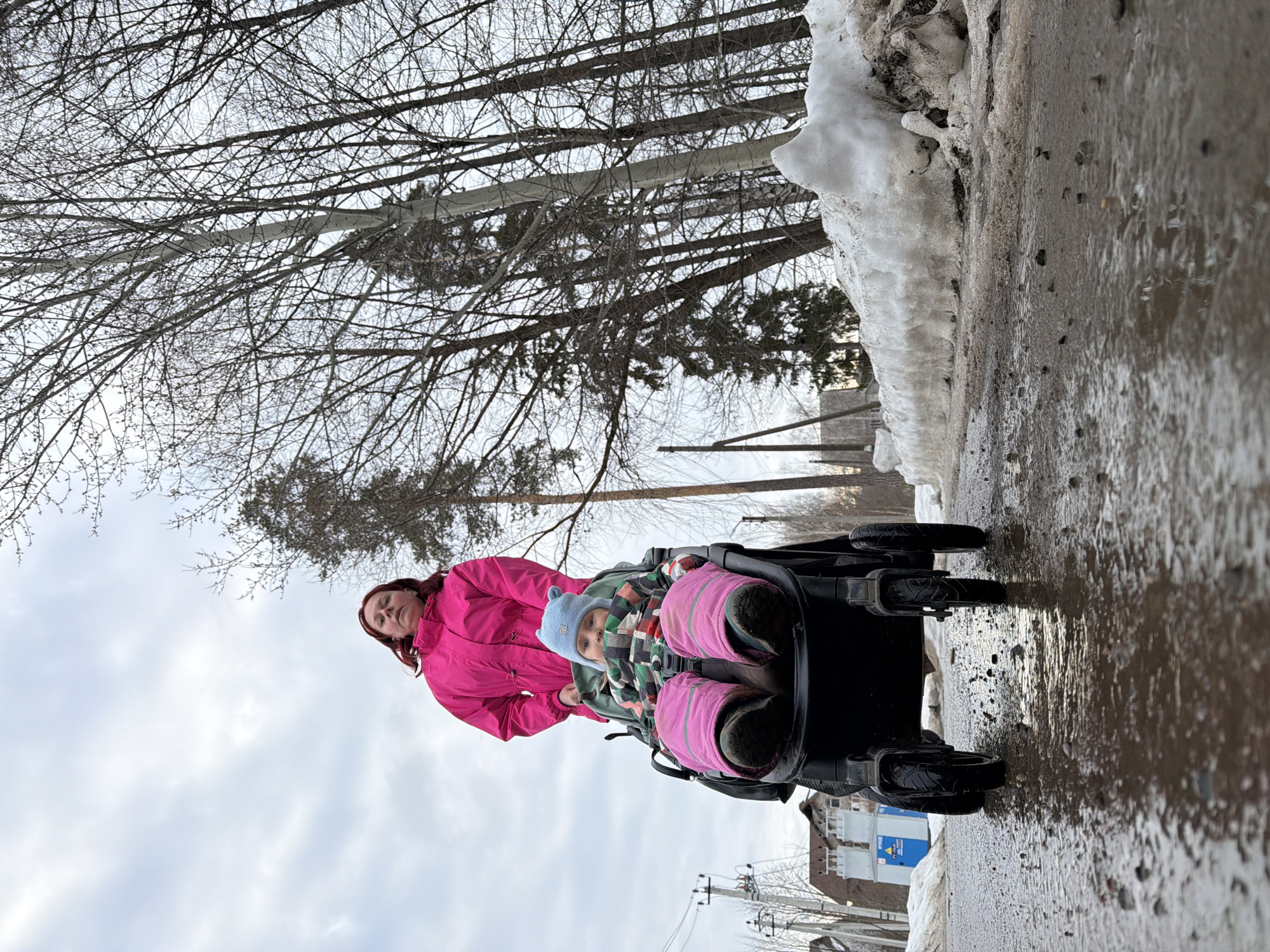 This image depicts an outdoor urban environment in Chaikovsky, Russia, during what appears to be late winter or early spring. The scene is captured from a low-angle perspective, emphasizing the foreground.
An adult female, with reddish-brown hair, is visible from the waist up, wearing a bright pink long-sleeved jacket. She is positioned behind and appears to be pushing a dark-colored stroller. Seated within the stroller is an infant or toddler, wearing a light blue knitted hat with ear-like protrusions, a multi-colored plaid garment, and pink insulated lower body covering. The infant faces forward, directly towards the camera.
The stroller is actively traversing a wet, muddy, and gravel-strewn path, with numerous puddles reflecting the overcast sky. Its four wheels are visible, with the front wheels prominent in the immediate foreground. To the right of the path, a bank of melting snow and slush is present.
The background consists primarily of numerous bare deciduous trees with thin branches and some evergreen trees. On the left side of the frame, multiple wooden utility poles with overhead wires are visible. Further to the left in the background, a multi-story building with a brown facade and a sloped roof is partially obscured, along with an outdoor electrical substation enclosure featuring blue and grey transformer-like components. The sky is uniformly overcast and grey.