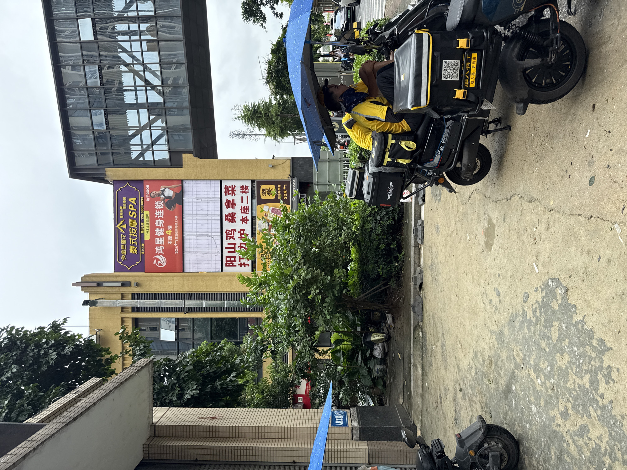 A street scene in Haizhu District, China features a delivery person on a scooter under two blue umbrellas. The delivery person, wearing a yellow shirt and a dark bandana, is seated on the scooter, which has multiple black delivery boxes with yellow accents. In the background, a building with a large yellow advertisement is visible, along with a modern glass skybridge. The ground is paved and appears damp. Other vehicles and foliage are also present in the scene.