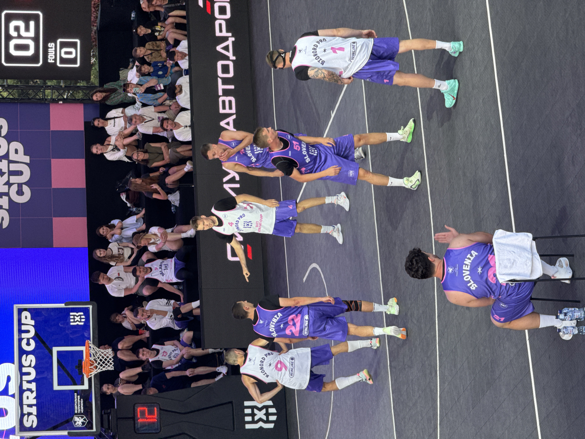 The image depicts an outdoor basketball game at the Sirius Cup in Sochi, Russia. The court is black with white lines, and a large scoreboard displays "02" for fouls and "0" for fouls. Spectators are seated in tiered bleachers behind the court. On the court, several basketball players are visible. One player in a white jersey with the number "9" and "IONORD PRO" on it is facing away from the camera. Another player in a purple jersey with "SLOVENIA" and the number "22" is also visible. A third player in a white jersey with "IONORD PRO" and the number "4" is pointing. Another player in a purple jersey with "SLOVENIA" and the number "51" is looking down. A player in a white jersey with "IONORD PRO" and the number "1" is also present. In the foreground, a person in a purple jersey with "SLOVENIA" and the number "22" is sitting in a chair, facing away from the camera. The scene suggests a competitive basketball match in progress.