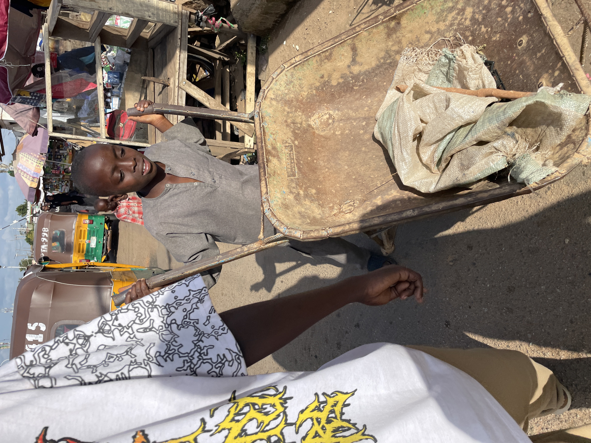 In Gura Top, Nigeria, a young boy in a grey shirt is pushing a rusty wheelbarrow filled with a soiled cloth bag. He is standing on an asphalt road next to a market. In the foreground, the lower half of a person wearing a white t-shirt with a yellow design and tan pants is visible, also on the road. Behind the boy, a man wearing a red and white plaid shirt is walking. In the background, a brown and yellow tricycle with the license plate "TA-998" is parked. The market features various stalls with goods displayed, including textiles. The scene is illuminated by sunlight, casting shadows on the ground.