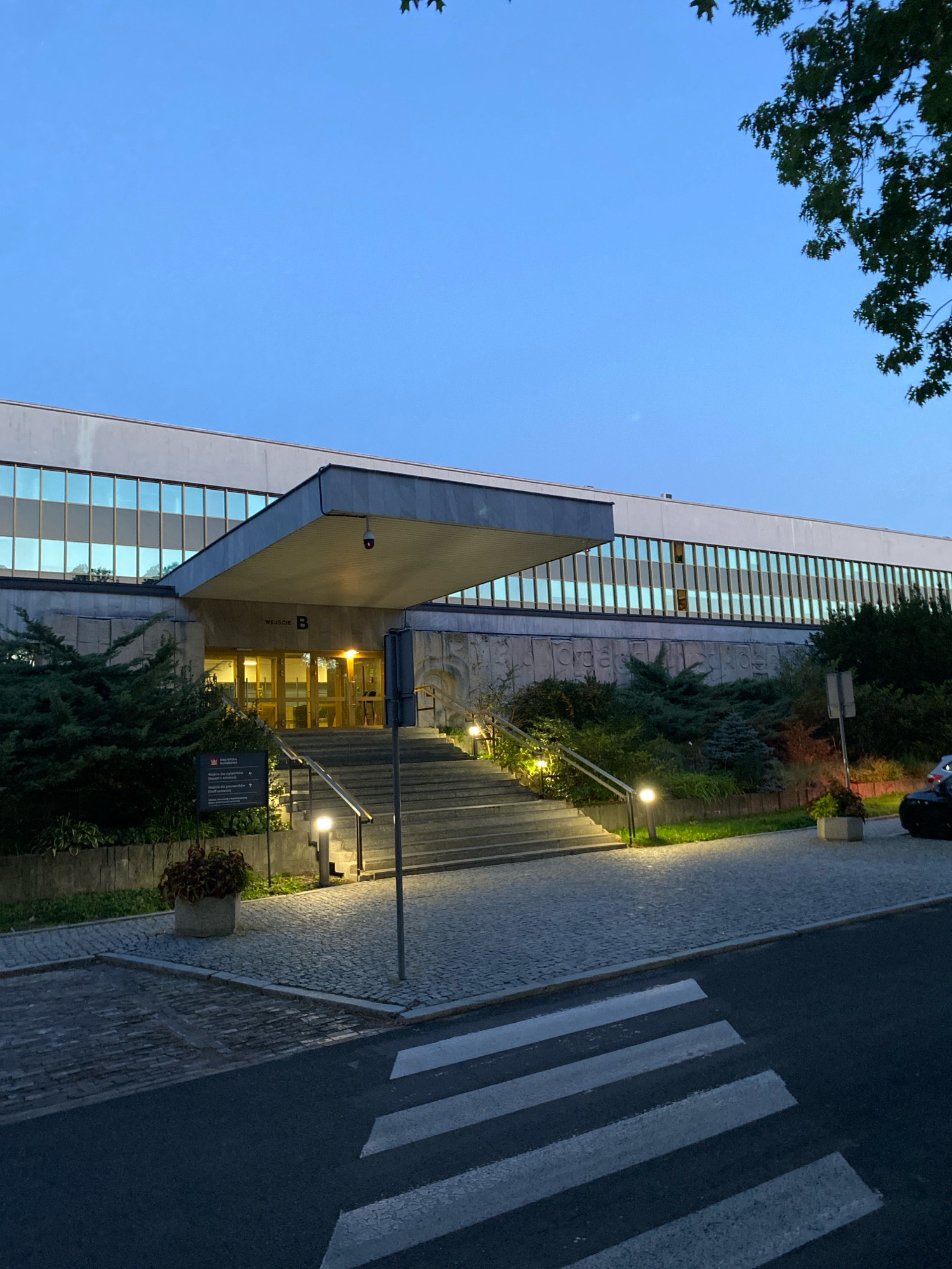 The image depicts the entrance to Building B at the National Library of Poland in Warsaw. The library's modern architecture features a long facade with a band of large windows reflecting the twilight sky. A wide, covered entrance leads to glass doors, and a set of stairs ascends to this entrance. The surrounding area is landscaped with evergreen bushes and illuminated by pathway lights. A crosswalk is visible in the foreground on a dark asphalt road. The sky is a deep blue, indicating early evening. There are no people visible in the image.