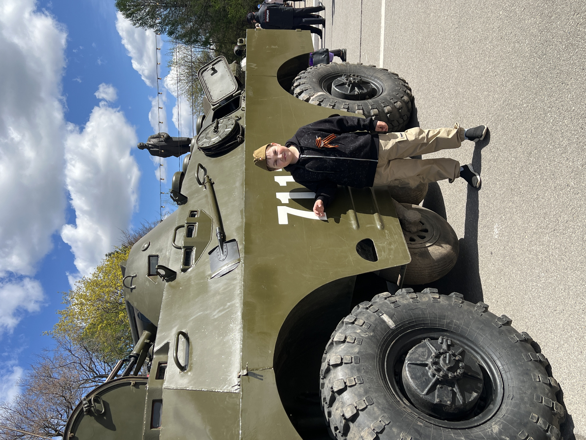 A child is seen posing on a green armored vehicle in Cherepovets, Russia. The boy wears a dark jacket with an orange and black ribbon, khaki-colored pants, and a military-style cap. The vehicle has the number "117" painted on its side. The surroundings include trees, a cloudy sky, and a paved surface. In the background, other individuals are visible, including those in police uniforms. A statue-like figure is also positioned on the armored vehicle.