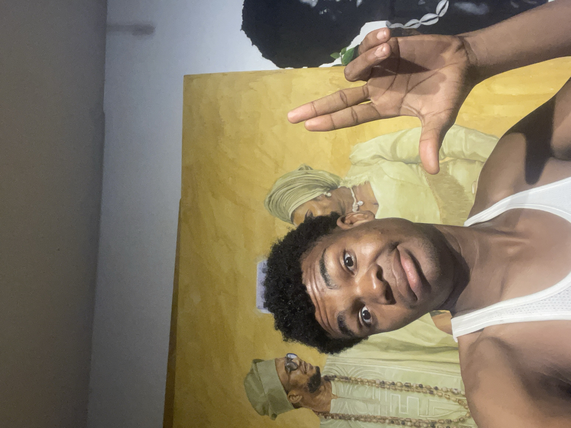 A young Black male in a white tank top is taking a selfie in front of a mural. The mural depicts three figures in traditional Nigerian attire, two women and one man, all wearing yellow and green garments. The man in the mural wears a green cap and beaded necklaces, while one of the women wears a green headwrap and pearl necklace. The background of the mural is a textured yellow. The young male in the foreground is making a gesture with his right hand, forming a "L" shape with his index and middle fingers and an "O" shape with his thumb and index finger. His facial expression is one of playful surprise, with wide eyes and an open mouth. The lighting suggests an indoor setting, possibly a gallery or cultural center in Ado, Nigeria.