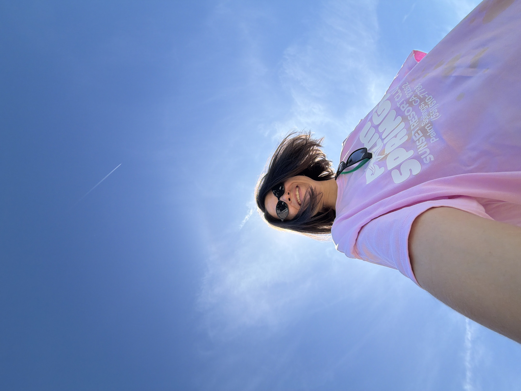 A low-angle shot captures a smiling person against a vibrant blue sky, positioned in what is stated as Kubasi, Montenegro. The subject, a person with dark hair, is seen from the chest up, looking down at the camera with a wide, happy smile. They are wearing dark sunglasses on their face, and another pair of black sunglasses with green earpieces is hooked onto the collar of their light pink t-shirt. Bright sunlight creates a halo effect around their head and shoulders, indicating a high sun.
The overwhelming background is a clear, bright blue sky, signifying sunny weather. A distinct white contrail from an airplane is visible in the upper left quadrant of the sky, and some very faint, wispy clouds are present, especially near the bright glare of the sun.
The person's cheerful expression suggests happiness or enjoyment.
Visible text on the pink t-shirt, oriented for the camera's perspective, includes "SUNSET RESORT CLU" and "Palm Springs, CA 92262", along with a phone number "(551) 610-7788". Larger, stylized text partially reads "PALM SPRING". These textual details reference Palm Springs, CA, which contradicts the given location context of Kubasi, Montenegro, as no visual cues within the image support the Montenegro location. The time of day is clearly bright daylight, consistent with the sunny conditions.