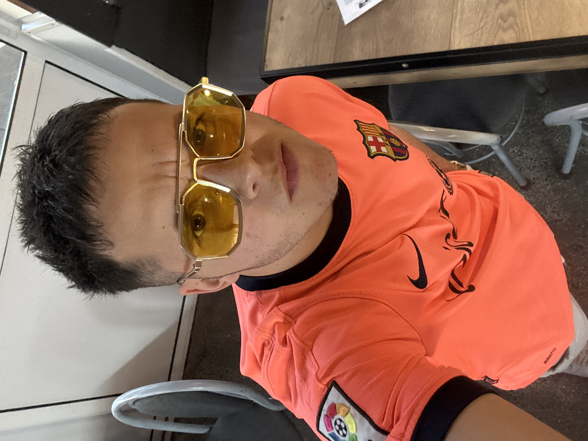 A man wearing a bright orange FC Barcelona soccer jersey and yellow-tinted sunglasses is sitting at a table. The jersey features the FC Barcelona logo on the upper left chest and the La Liga logo on the lower left hem. The man is indoors, and part of a gray chair and a white tiled wall are visible in the background. The location is Kostinbrod, Bulgaria.