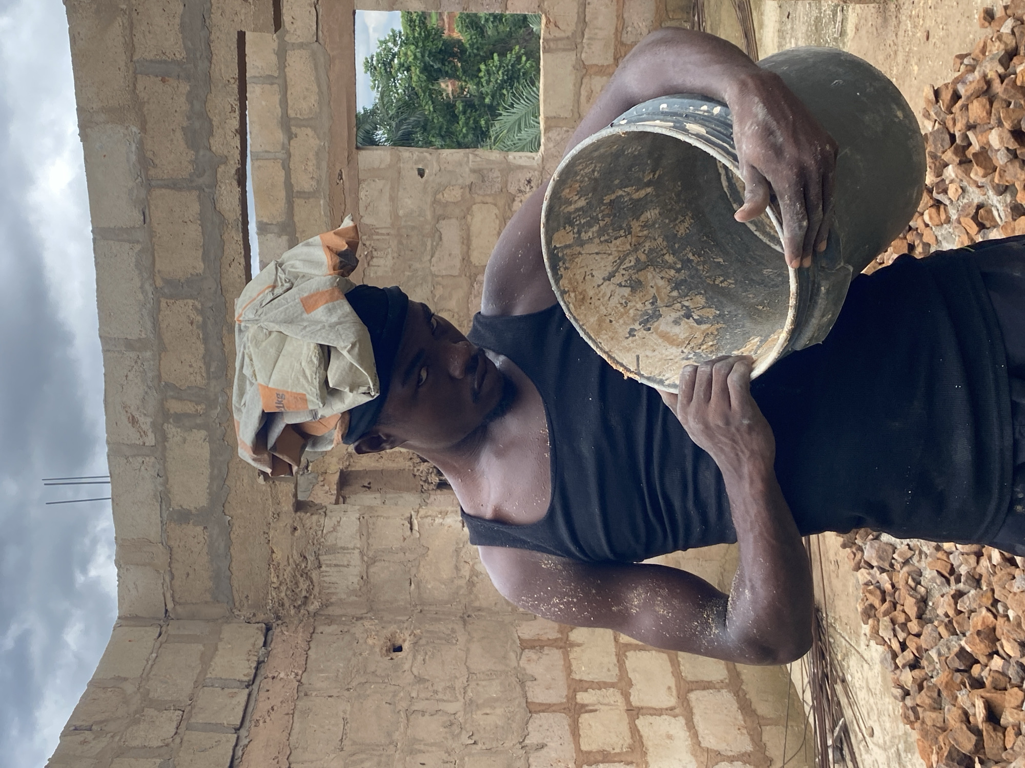 A man in Ufu Esioni, Nigeria, wearing a black tank top and a head covering made of fabric, is holding a large, dirty bucket. He appears to be inside an unfinished building constructed of tan-colored blocks. The building has an open window showing green trees outside. Construction debris is scattered on the ground.