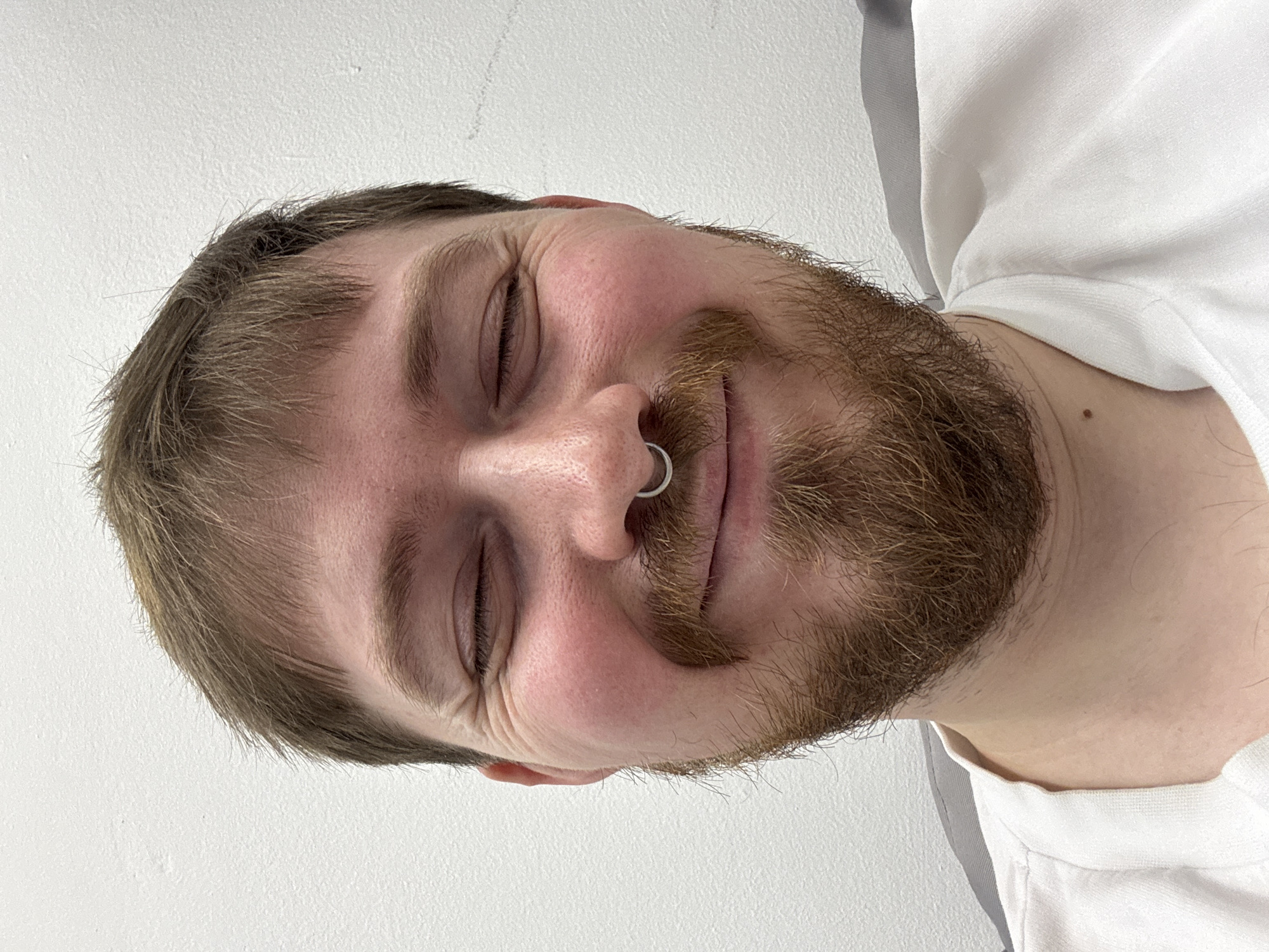 A close-up, rotated portrait of a bearded man with a fair complexion. He has short brown hair and a brown beard with a prominent mustache. He has a silver septum piercing in his nose and his eyes are closed. He appears to be smiling slightly. He is wearing a white t-shirt. The background is a plain, light-colored wall. There is no information present to establish the location as the city of Chaikovsky, Russia.