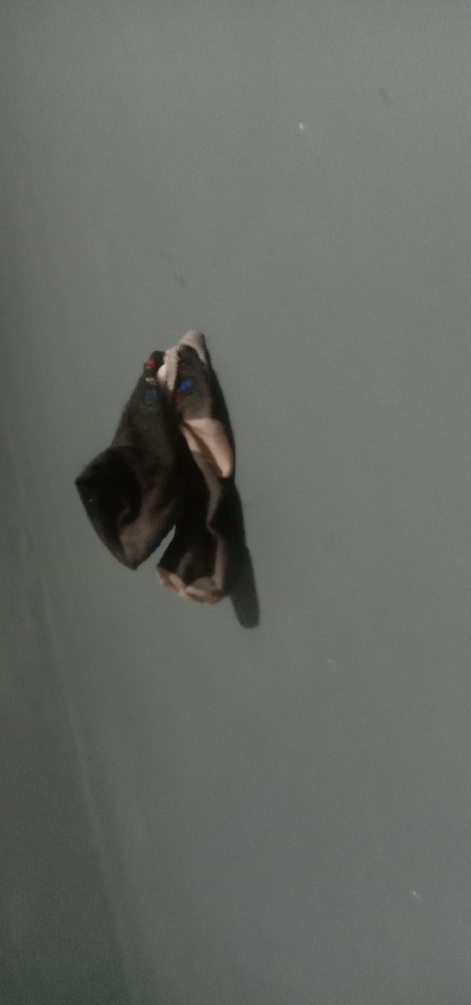 The image depicts two black gloves, possibly leather, that appear to be wet or soiled, resting on a smooth, gray, non-reflective surface. The gloves are positioned together, with one slightly overlapping the other. Small, colored markings, possibly blue and red, are visible on the surface of the gloves. The lighting is dim, casting subtle shadows. The background is a uniform, dark gray, suggesting a nondescript indoor or sheltered outdoor environment. There is no information in the image that allows for the determination of its location in Kendari, Indonesia.