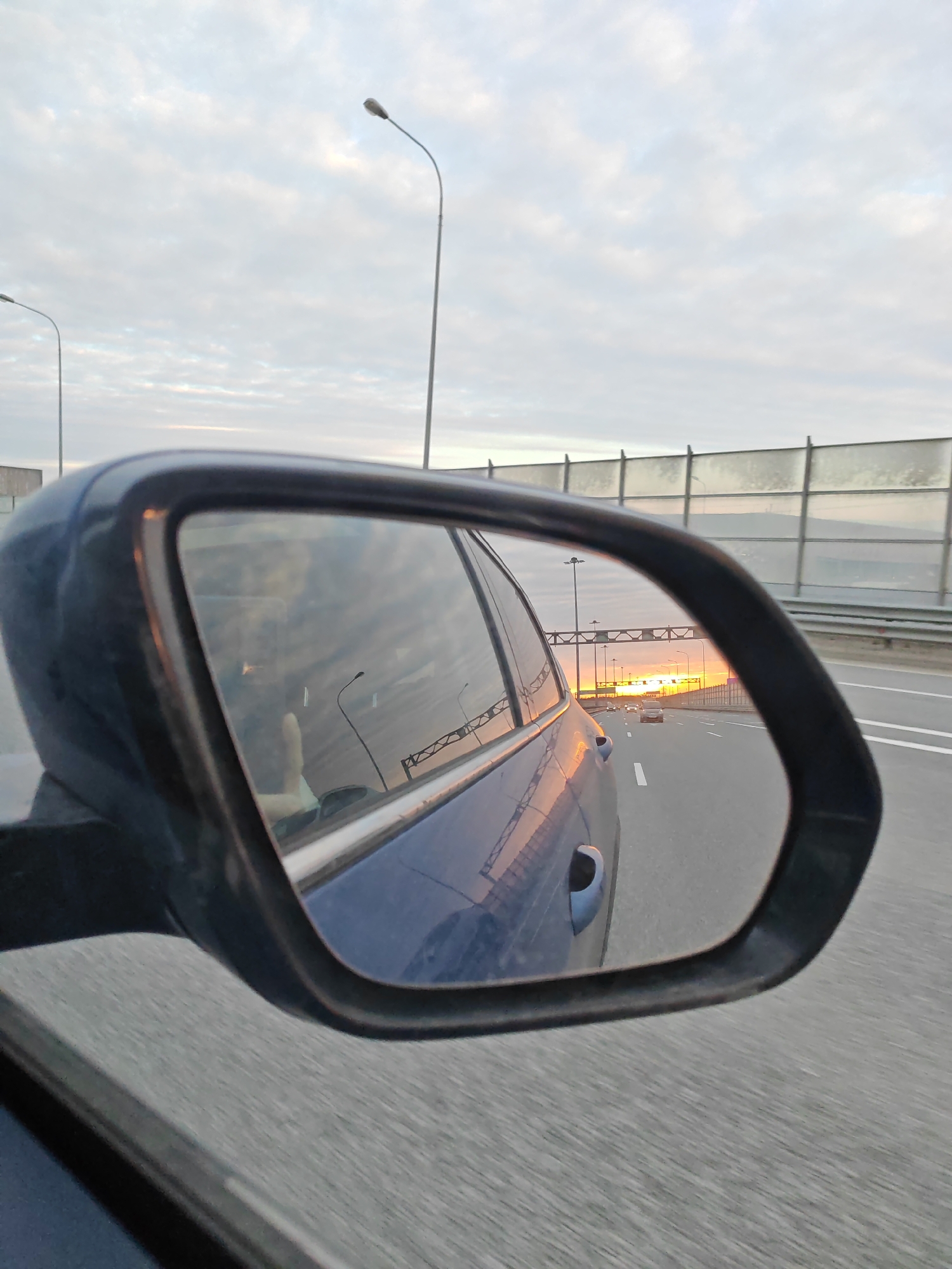 A daytime shot, taken from a moving vehicle on a highway near Poroshkino, Russia, showcases a side mirror reflecting the road behind. The reflection reveals a multi-lane highway, featuring at least two other vehicles traveling in the same direction. A sunset is visible in the distance, casting a warm glow on the road and adjacent sound barriers. The sky is overcast with light clouds. The image appears to be taken in motion, as evidenced by the blurred road surface. The side mirror's frame and the car's body it is attached to are also visible in the foreground. Streetlights line the side of the highway.