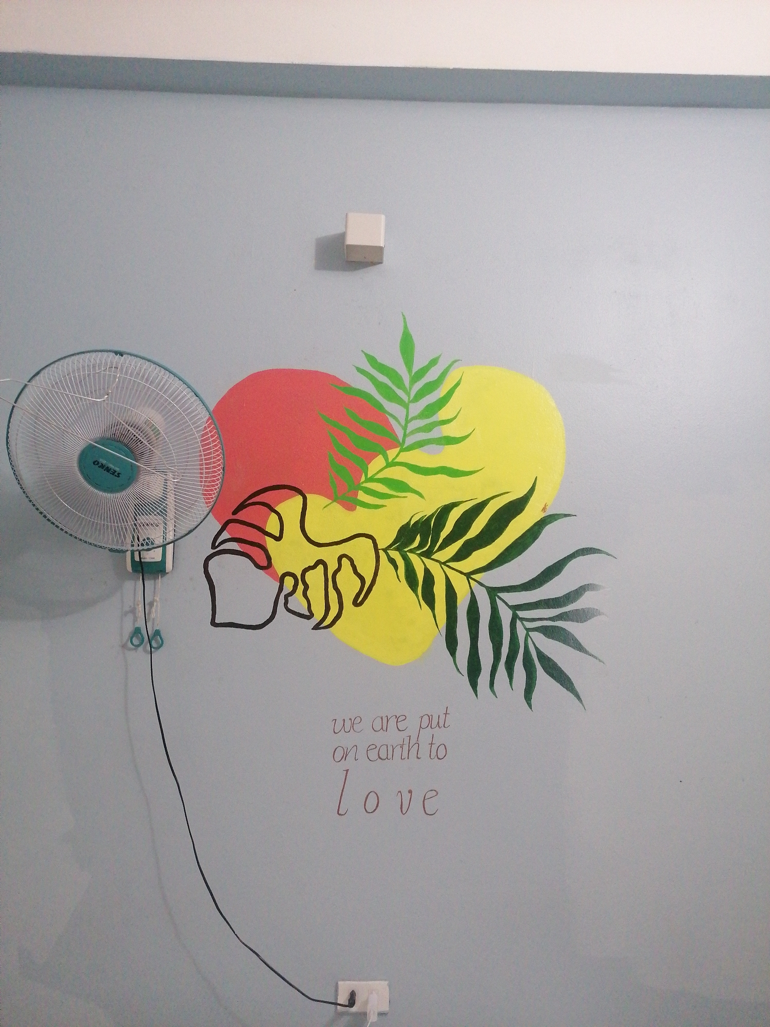 The image depicts a section of a wall in an indoor setting. The wall is painted light blue and has a decorative design featuring a red and yellow circular shape with painted green leaves. A black outline drawing of an abstract object is positioned on the colored shapes. Below the artwork, the text "we are put on earth to love" is written in brown font.
A wall-mounted fan is visible on the left side of the frame. The fan has a green center and a white grill. An electrical cord extends from the fan down to a wall outlet near the bottom of the frame. A white electrical box is located near the top center of the frame.
There is no indication of Bai Chay Ward, Vietnam.