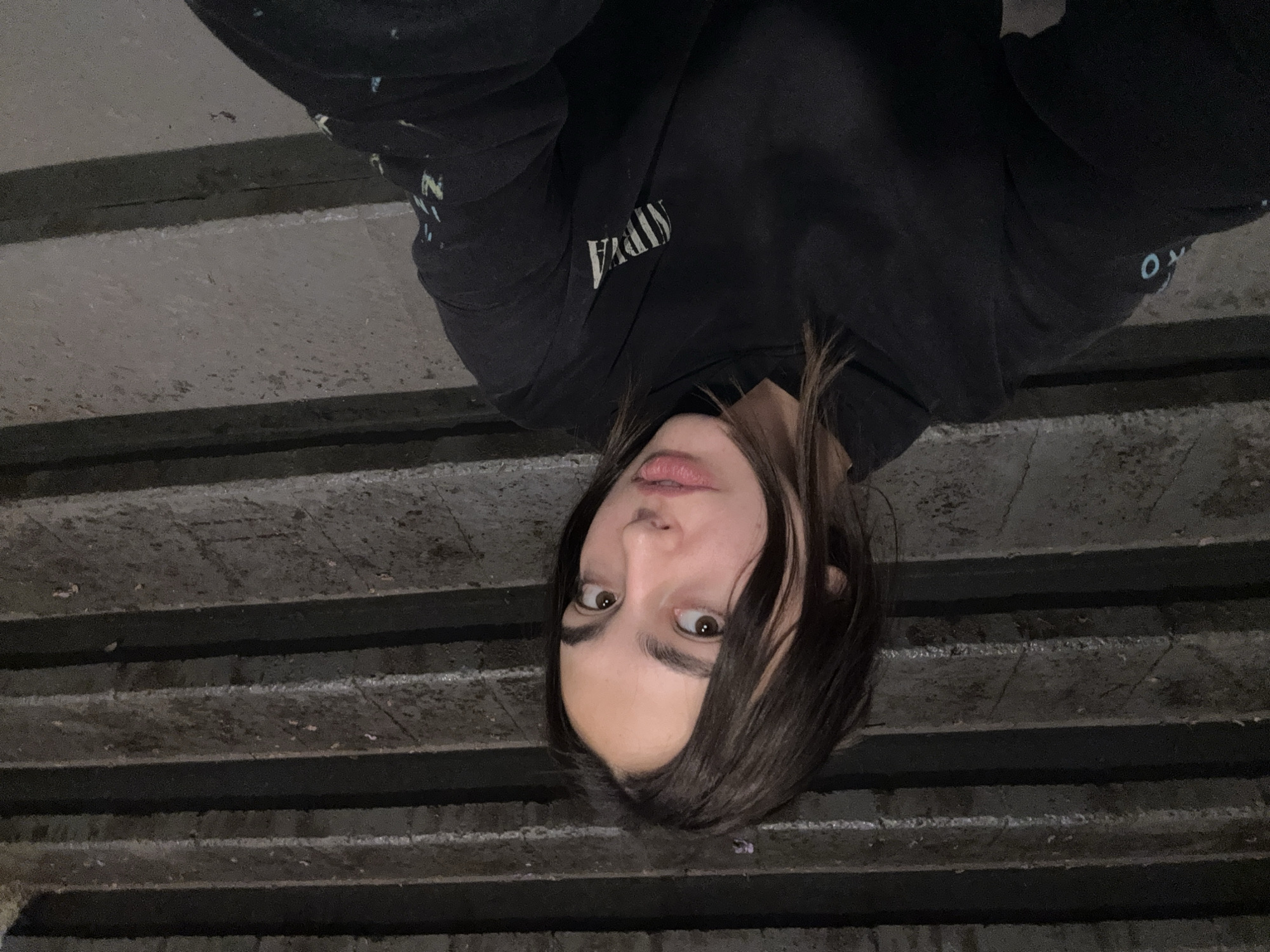 This overhead selfie, rotated 90 degrees clockwise, features a young person with dark, medium-length hair looking directly at the camera. The subject has expressive brown eyes and slightly parted lips, maintaining a neutral to slightly curious gaze. They are wearing a dark-colored hoodie, with the word "NIRVANA" clearly visible in white, upside-down text across the chest. Indistinct light-colored designs or text are also present on both sleeves.
The background consists of a series of parallel, dark, and textured horizontal strips, strongly resembling concrete steps or risers viewed from above. The overall lighting is dim, suggesting either an evening or nighttime setting, or an indoor environment with low illumination. The unique perspective indicates the individual is taking the photo from an elevated position, looking down at themselves. While the image is provided with a location context of Mexico City, Mexico, there are no specific visual cues within the photograph itself to confirm this geographical detail. The atmosphere conveyed is quiet and somewhat intimate due to the close-up, self-portrait nature of the shot.