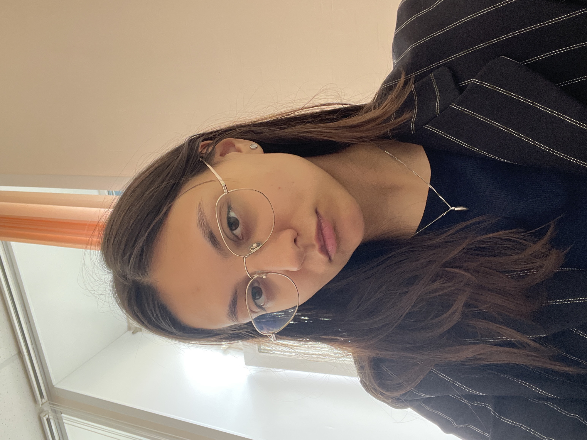 A woman with long brown hair, wearing glasses and a black pinstripe blazer, is pictured in a close-up, slightly angled, portrait. She is also wearing a dark top and a silver necklace. The background includes a white wall and what appears to be a window or window frame. There is not enough information to ascertain a specific location in the city of Yekaterinburg, Russia.