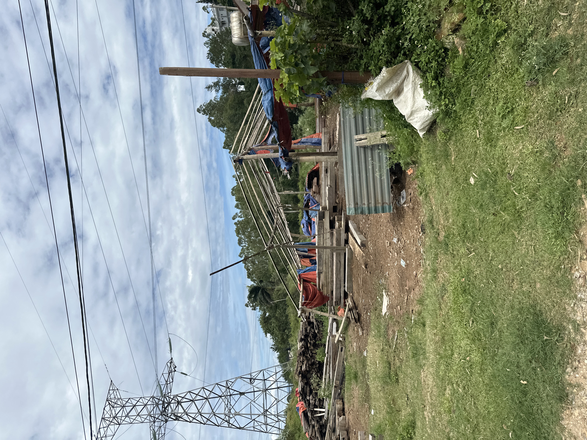 In Bắc Yên, Vietnam, a construction site is visible with a large electrical tower dominating the left side of the frame. The site contains a partially constructed wooden frame structure covered with blue and red tarps, suggesting temporary shelter or roofing. Piles of lumber and debris are scattered around the area. In the foreground, green grass covers the ground, with a white bag and some scattered trash. The sky is cloudy, with a mix of blue sky and white clouds. The overall impression is one of an active but somewhat unkempt construction area.
