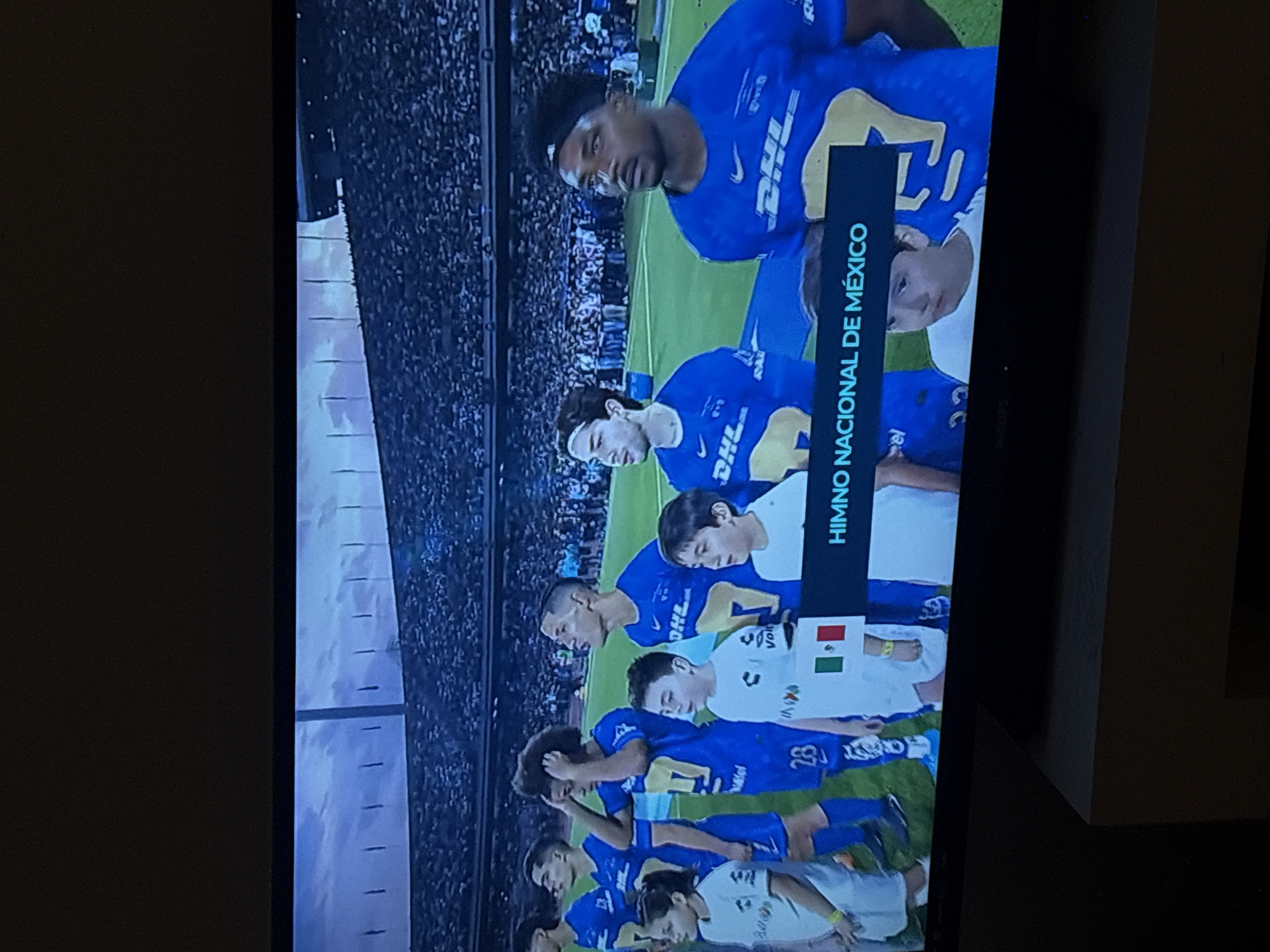 A television displays a scene of a soccer team wearing blue uniforms with the DHL logo standing on a field, presumably in Mexico City, Mexico. The text on the screen reads "HIMNO NACIONAL DE MEXICO," indicating the players are about to sing the Mexican national anthem. Some players are wearing white shirts with a small Mexican flag. The scene is likely taking place in a stadium with a large crowd in the background.