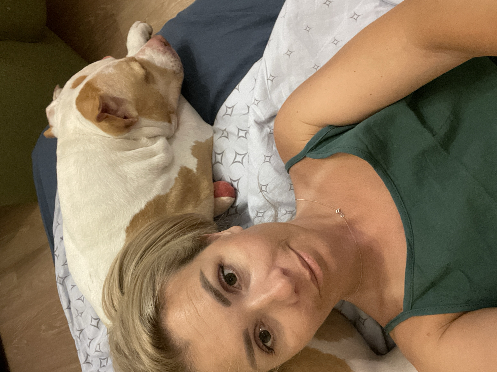 This is a selfie taken from a high angle, looking down at the subject. The subject is a woman with blonde hair, wearing a dark green tank top and a delicate silver necklace. She is lying down on a bed, with her head tilted. Next to her, also lying down, is a dog. The dog is predominantly white with tan markings around its head and ears, and appears to be sleeping. The bedding has a subtle geometric pattern of outlined diamonds. The scene appears to be indoors, likely a bedroom, given the presence of a bed and bedding. The lighting suggests it might be daytime, but the specific time of day is not discernible. The woman has a relaxed expression. There is no visible text in the image.