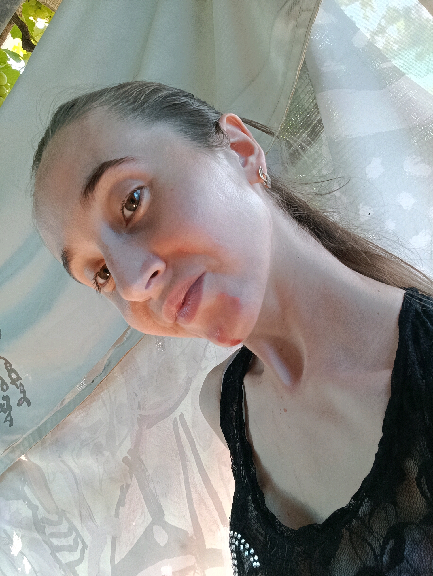 The image shows a close-up, low-angle selfie of a young woman, appearing to be outdoors in an area with vegetation and a draped fabric. The woman has light brown hair pulled back, and is wearing a black lace top. She has a notable amount of red makeup or paint applied to her lower face, particularly around her mouth and chin, giving the appearance of blood. Her skin is fair, and she has brown eyes and light colored eyebrows. She is wearing a small hoop earring in her left ear. The background consists of a light-colored, semi-transparent fabric that is draped and possibly attached to a structure, with some leafy green foliage visible in the upper left corner. The lighting suggests it is daytime. The setting does not provide any definitive visual cues to ascertain the specific location within Vypasne, Ukraine.