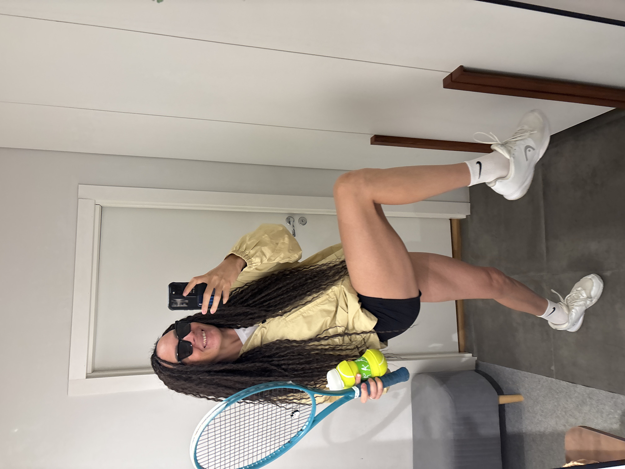 A person is captured in a mirror selfie, standing indoors in what appears to be a room or hallway. The individual, likely female, has long, dark, curly hair and is wearing black sunglasses that partially obscure a wide smile. She is dressed in a light yellow, long-sleeved jacket over a white top, black athletic shorts, white ankle socks with a black Nike swoosh, and white sneakers featuring the "HEAD" logo on the side.
She holds a light blue tennis racket in her left hand. Her right hand, holding a black smartphone to take the selfie, also grasps two yellow-green tennis balls, one with a partially visible "WILSON" logo. The person's stance and equipment suggest an association with tennis.
The background consists of light-colored walls, a white doorframe on the left, and a white ceiling with subtle horizontal paneling. A dark brown wooden shelf or beam is visible on the right wall. The floor is a dark gray, possibly concrete or textured surface, with a gray upholstered bench or ottoman partially visible in the lower right corner. The scene depicts a casual, self-portrait moment in an indoor setting in Chaikovsky, Russia.