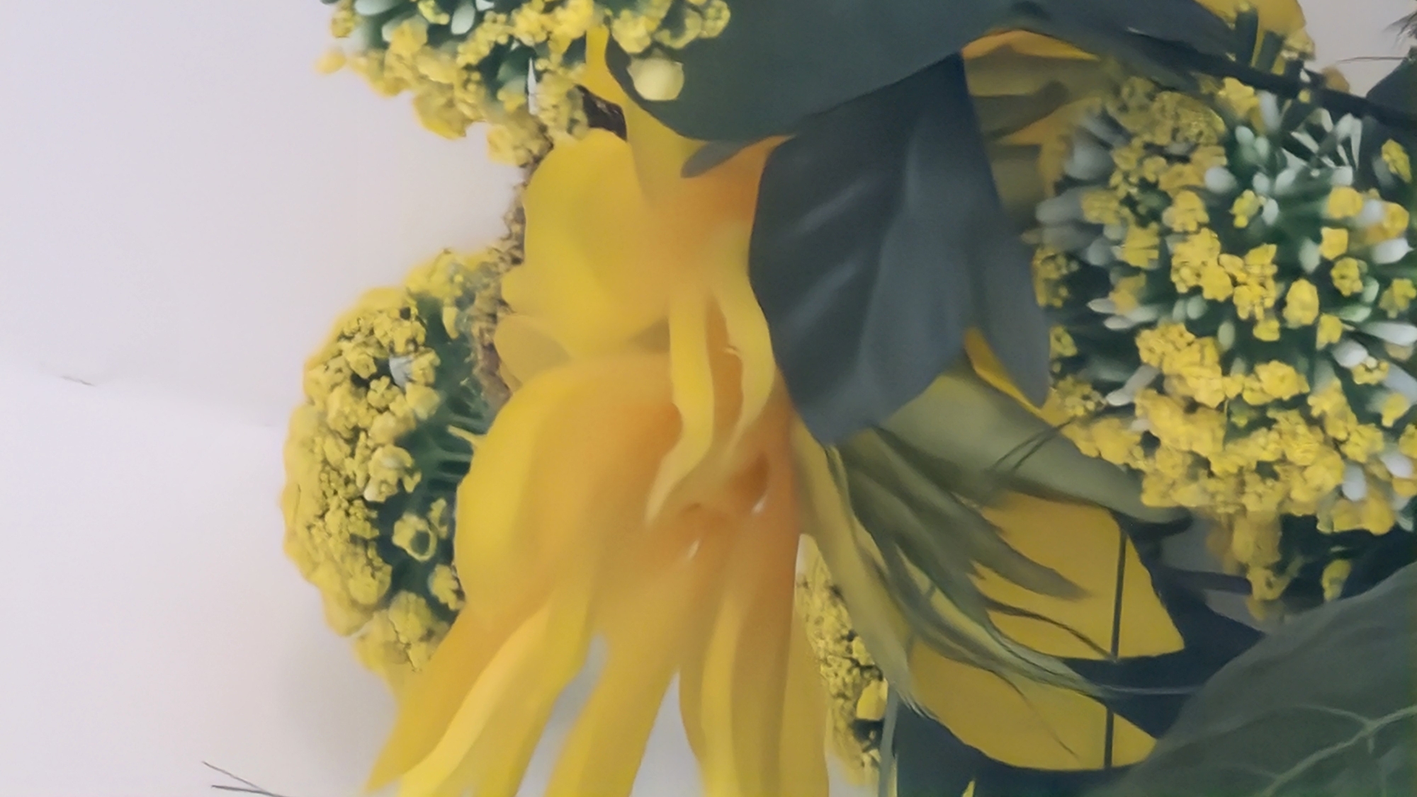 The image shows a close-up of a yellow floral arrangement against a white background. The arrangement includes a large, multi-petaled yellow flower, possibly a sunflower or dahlia, surrounded by smaller clusters of yellow and white flowers, and dark green leaves. The composition is dense, filling the frame. The location in the city of Lincoln, United Kingdom is not discernible from the image. There are no people, subjects, actions, or interactions visible. The only objects present are the floral arrangement and the background.