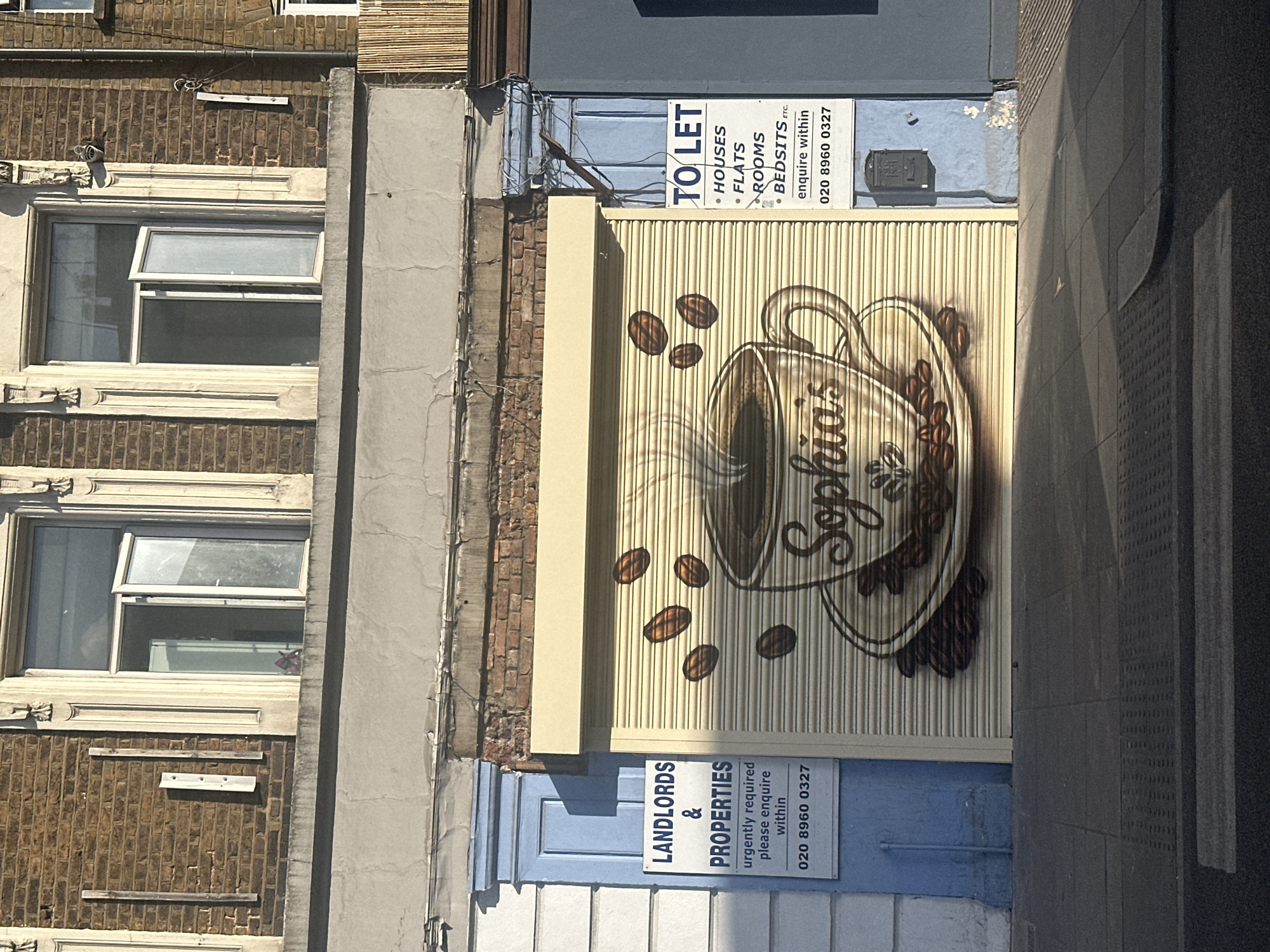 This photograph shows the facade of a building in London, United Kingdom. The building has a combination of architectural styles, including brickwork, render, and corrugated metal shutters. A mural of a coffee cup with the text "Sophia's" is painted on one of the shutters. Signs reading "TO LET" and "LANDLORDS & PROPERTIES" are posted on the building, each listing a phone number. A window is visible on the brick section of the facade. The shot is taken from street level, displaying a portion of the pavement.
