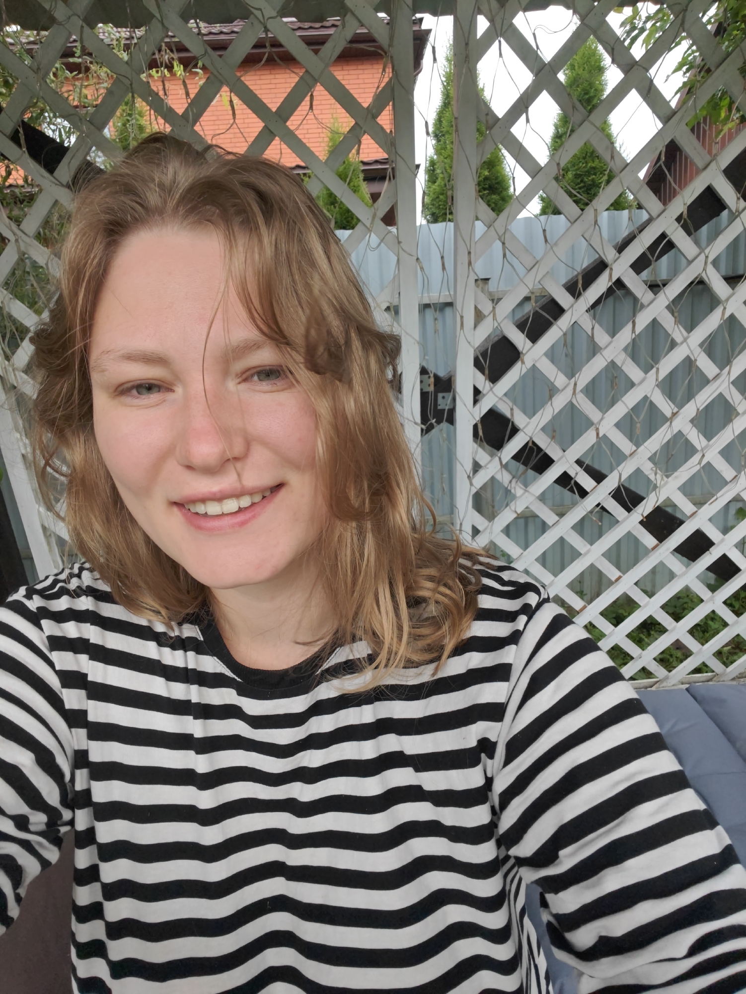 A young woman with blonde, shoulder-length hair smiles at the camera. She is wearing a black and white striped long-sleeved shirt. The background appears to be outdoors, behind a white lattice structure. A gray fence and green trees are visible behind the lattice. The image was taken in Koledino, Russia.