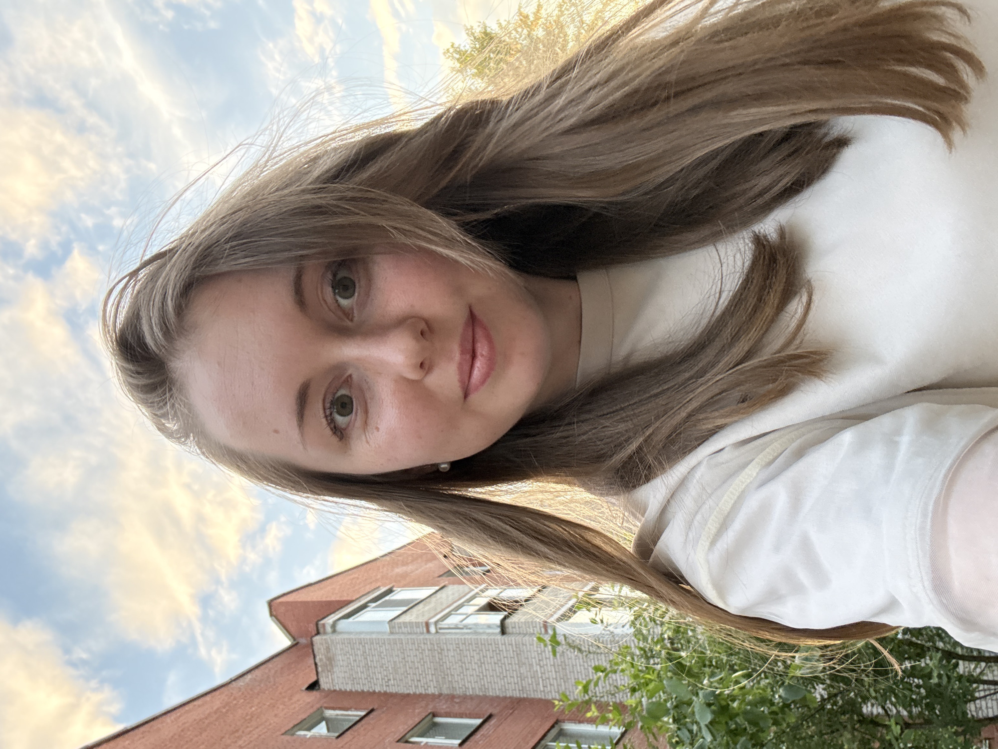 A young adult female is depicted in a close-up outdoor selfie. She has long, light brown hair, green or hazel eyes, a fair complexion with visible freckles, and a subtle smile, looking directly at the camera. A single small, round pearl earring is visible in her left ear. She is wearing a light-colored, possibly cream or off-white, long-sleeved top. The background features multi-story residential buildings constructed primarily of reddish-orange brick, with some sections having lighter, possibly white or grey, brick accents and balconies. Green foliage from bushes or trees is visible in the lower left and right corners of the frame. The sky above is partly cloudy, with the clouds illuminated by warm golden and pink tones, indicating either a sunset or sunrise. The light source appears to be behind and to the right of the subject, creating backlighting on her hair and the foliage. The scene is set in an urban or suburban residential area within the city of Unknown, Unknown.