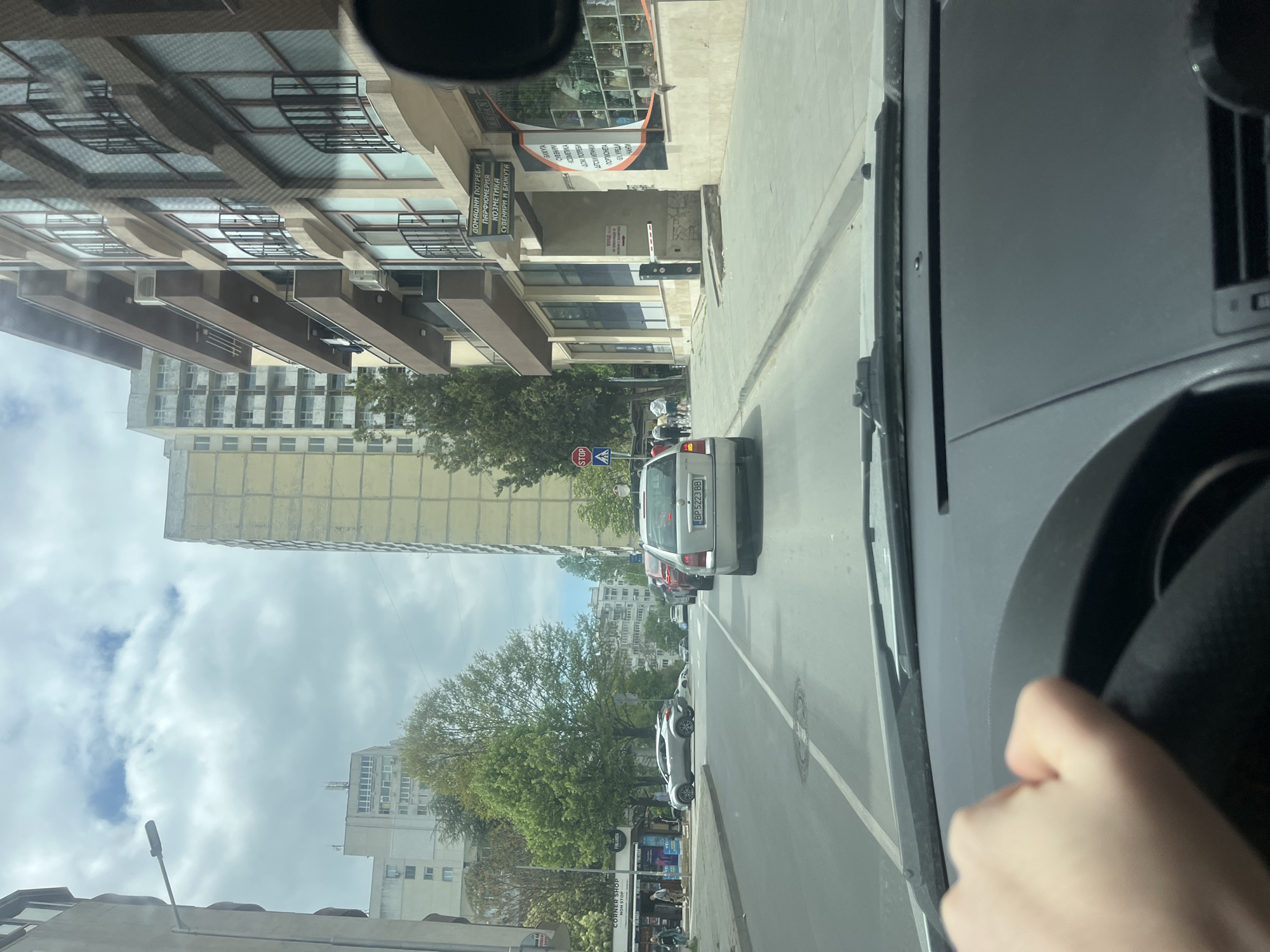 The image shows a street scene in Varna, Bulgaria, viewed from inside a vehicle. The driver's hand is visible on the steering wheel. The road ahead has a white car and a red car following behind, with a white car is parked on the right side of the road. The buildings along the street include a modern building with a shop named "Corner Shop," which sells cosmetics, perfumes, and souvenirs. There is a "STOP" sign visible near the corner shop. There are apartment buildings and trees visible further down the street. The sky is cloudy.