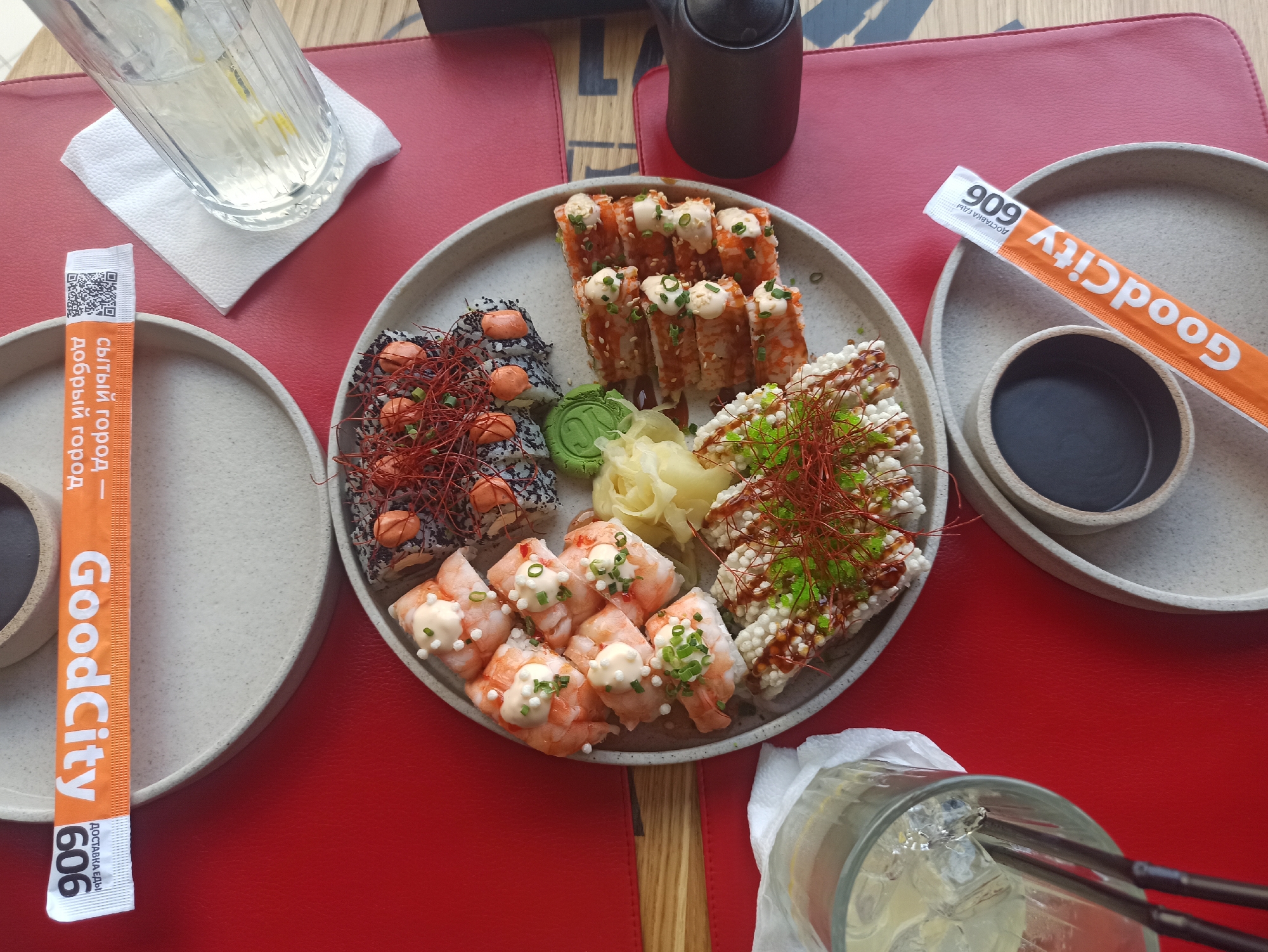 Here is a detailed description of the image:
The image shows a plate of sushi with several different types of rolls, along with ginger and wasabi. There are also two glasses of liquid with ice, possibly lemonade. Two plates are on the table, each with a small bowl containing dark liquid, probably soy sauce. There are orange wrappers for chopsticks labeled "GoodCity" in Russian, with the phrase "Сытый город - добрый город," which translates to "Satisfied city - kind city." The number "606" is also on the wrappers.
The setting is likely a restaurant, with a red tabletop and an underlying pattern of light-colored wood.