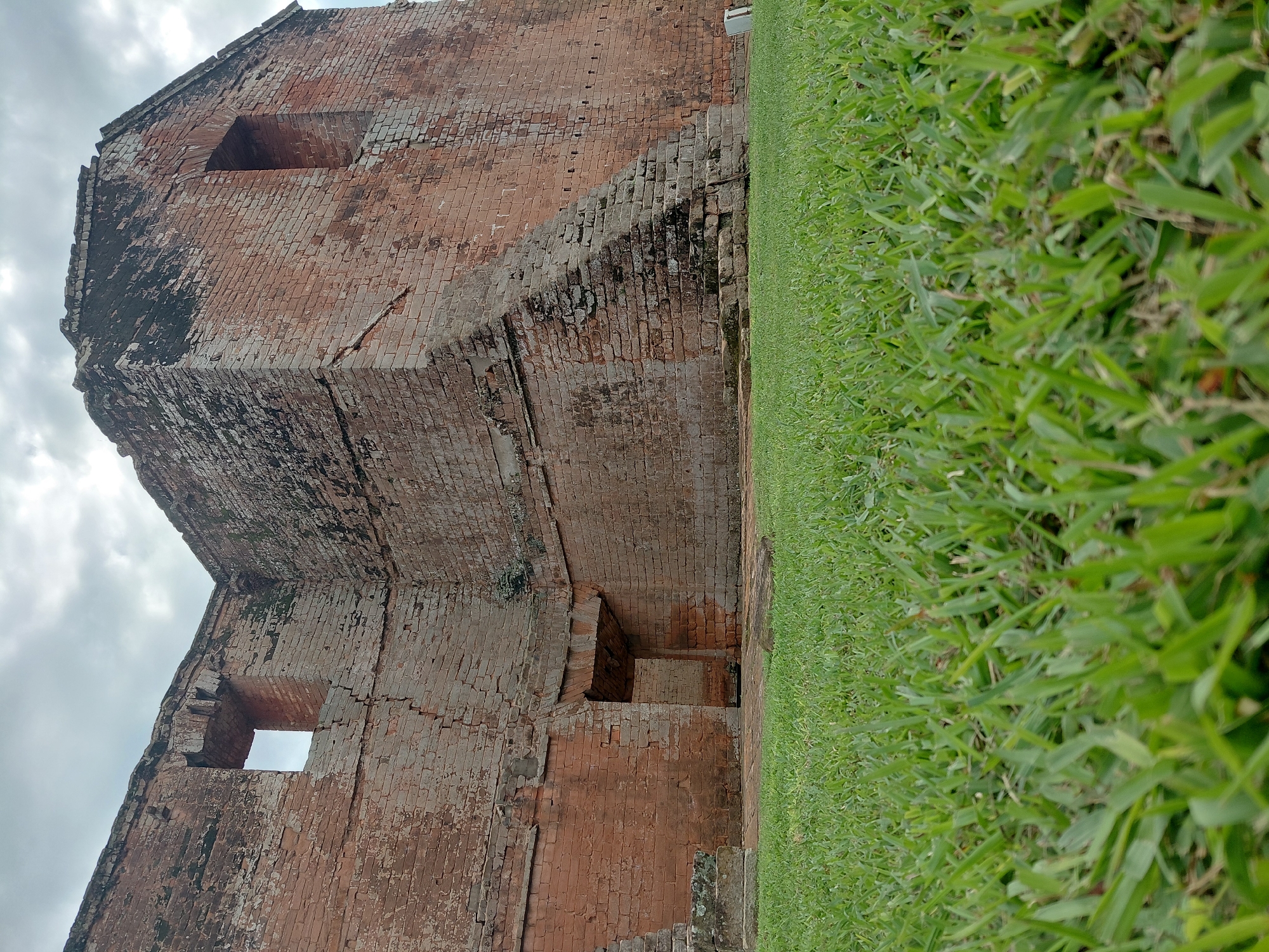 The image depicts the exterior of a weathered, red brick structure, likely a historical ruin. The building features arched openings and large rectangular window-like apertures. A prominent set of stone steps is built into the corner of the edifice. The foreground is dominated by a lush, green lawn, with the grass blades sharply in focus. The sky above is overcast with visible clouds. This structure is identified as the La Santísima Trinidad de Paraná mission in Trinidad, Paraguay.