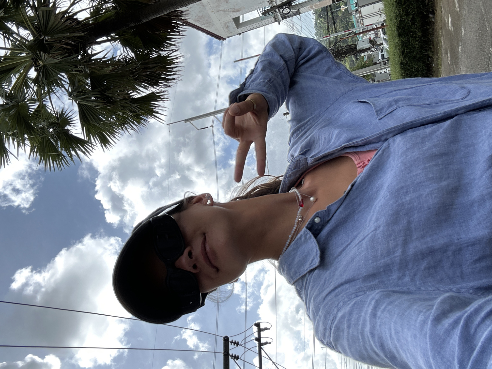 A person is taking a selfie outdoors with a cloudy sky. The person is wearing a blue collared shirt, sunglasses, a baseball cap, and jewelry. They are holding up two fingers in a peace sign. A palm tree is visible in the background, along with power lines, a building, and some vehicles. The scene appears to be in Kathu, Thailand.