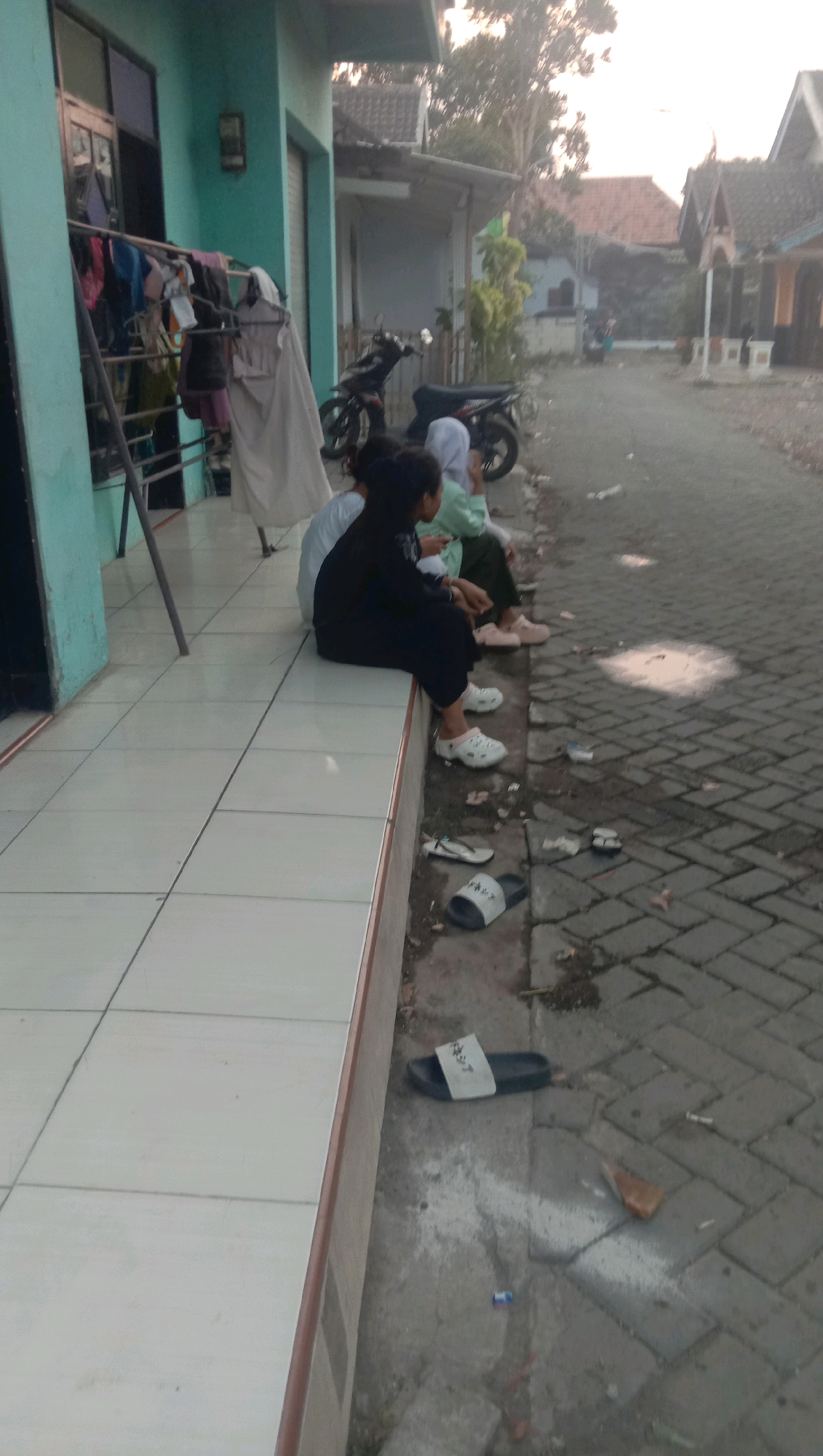 The image depicts a residential street scene in Karang Anyar, Indonesia. In the foreground, on a tiled porch, three individuals are seated. The person closest to the camera is wearing a black outfit and white clogs, facing away from the viewer. To their left, a person in a light green headscarf and white clogs is also seated. Further left, another individual in a white garment is partially visible. A clothesline with laundry is present on the porch to the left. A motorcycle is parked on the cobblestone pathway in the middle ground. The background shows houses with tiled roofs and trees. Scattered on the pathway are several pairs of sandals and flip-flops, along with some debris. The overall lighting suggests it is late afternoon or early morning.