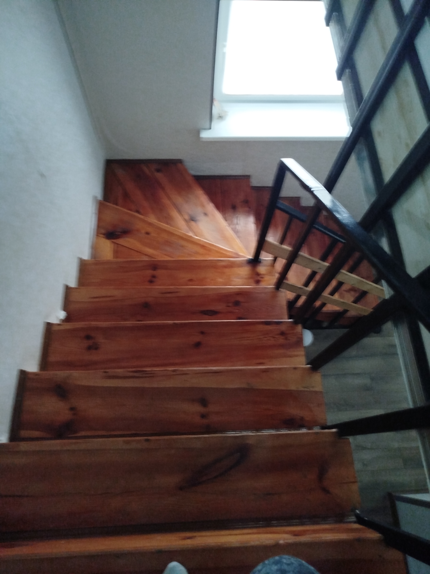 The image shows a wooden staircase with a glossy finish, leading upwards towards a brightly lit window or skylight. The staircase has visible wood grain and knots. There is a black metal railing on the right side of the stairs. The walls appear to be a light color, possibly painted. The floor beneath the stairs on the right is a muted tone, possibly wood or tile. The overall impression is an interior view of a staircase in a residential or public building. The image does not contain information to pinpoint the location to Крывасельскі сельскі Савет, Belarus.