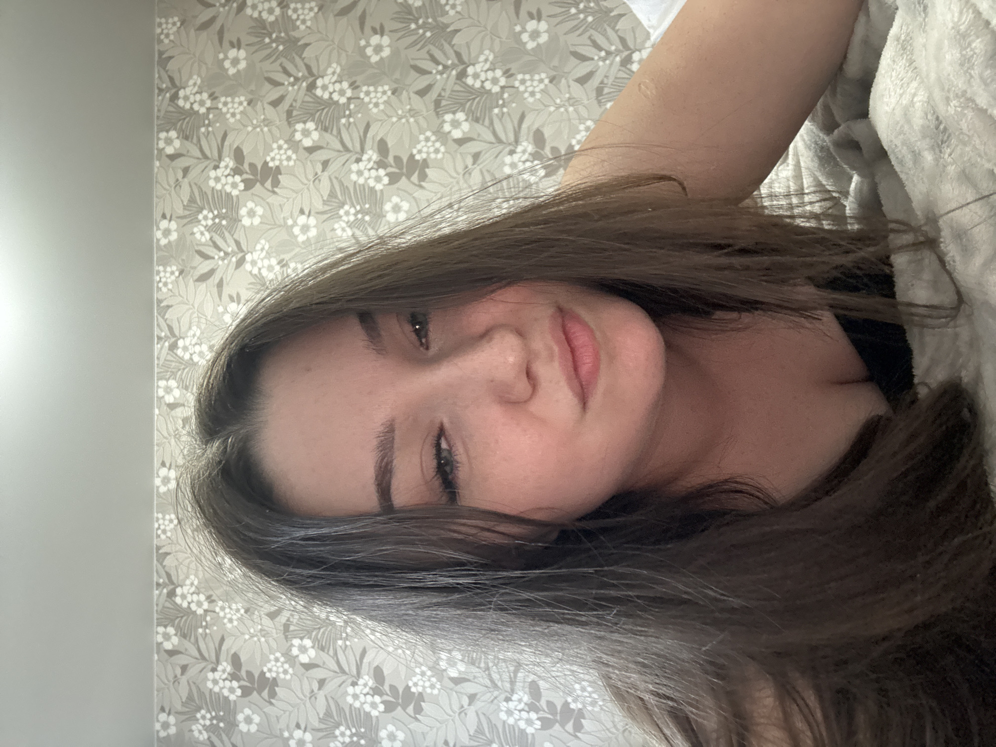 The media file shows a close-up, sideways selfie of a young woman lying down. She has long, dark brown hair that partially obscures her face. Her eyes are looking towards the camera, and her lips are slightly parted. She appears to be in a bedroom, as indicated by the floral wallpaper behind her and a soft, white blanket to her right. The lighting suggests it could be daytime, possibly indoors, as there are no strong shadows or indications of natural light sources. The woman's expression is relaxed and casual, suggesting a private moment at home. There is no visible text in the image. The context provided places the location as Zarechnaya (Gamovskoe S/P), Russia.