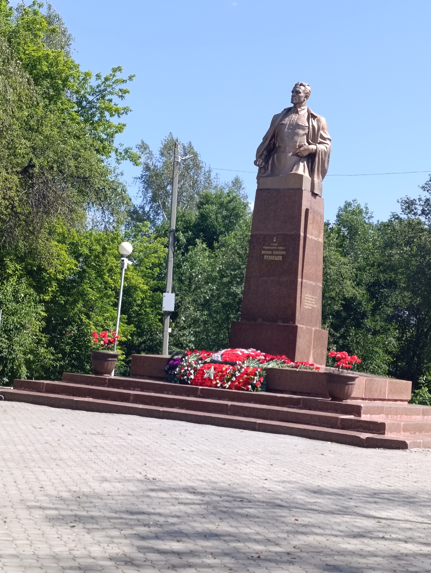 A statue of a man in military uniform stands atop a granite monument in Talitsa, Russia. The statue depicts a man with his left arm bent and resting on his chest, and his right arm hanging by his side. He is looking to his left. The monument is rectangular and made of red granite, with a base that has multiple tiers of stairs. There are wreaths of red flowers placed on the steps of the monument. The monument is surrounded by trees and a clear blue sky. The area in front of the monument is paved with gray interlocking pavers.