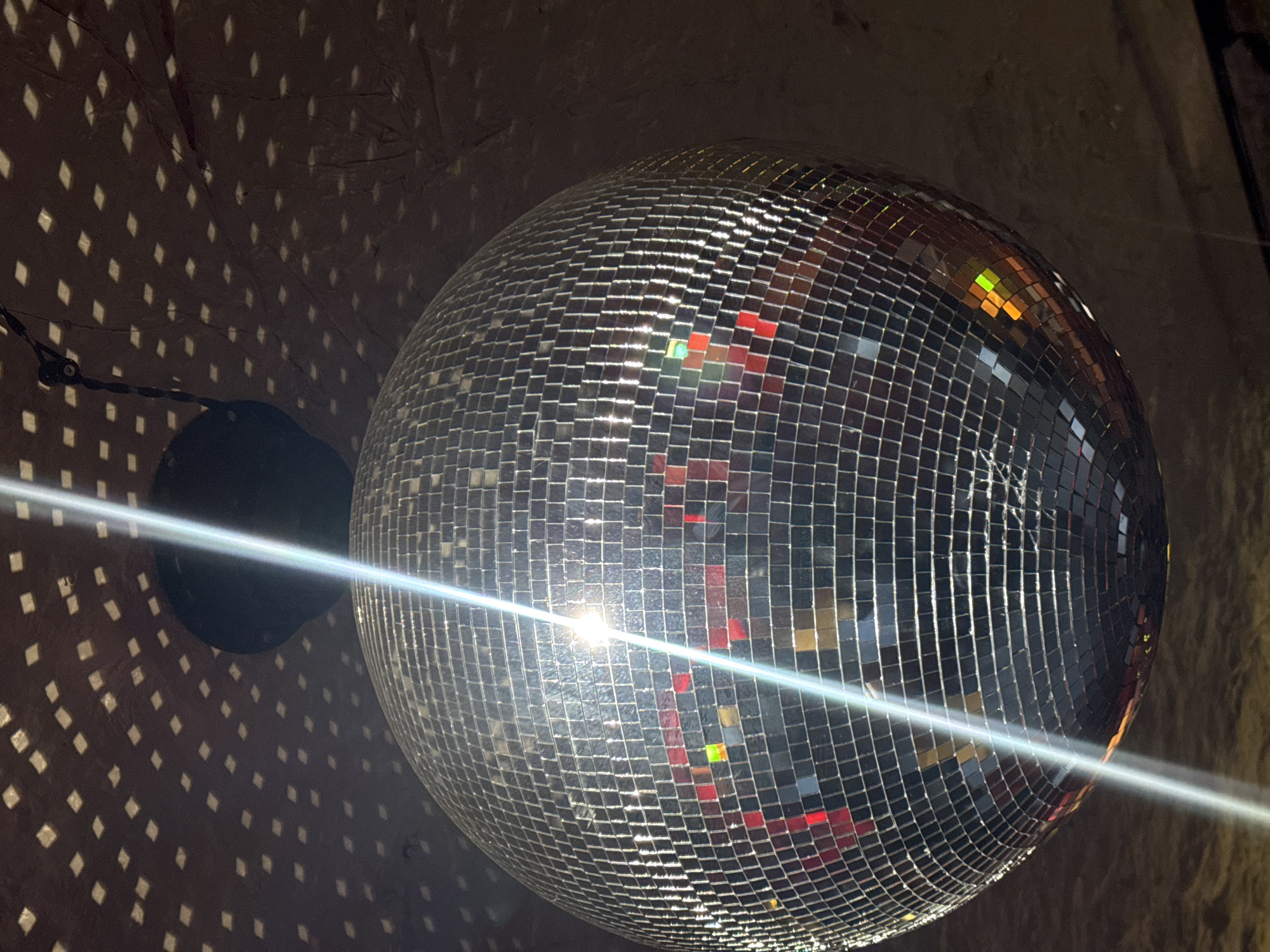 The image shows a disco ball hanging indoors. Light reflects off the disco ball, creating a pattern of small squares on the surrounding wall. A light source, likely a spotlight, shines directly onto the disco ball, creating a bright glare. The location of the image is unknown.