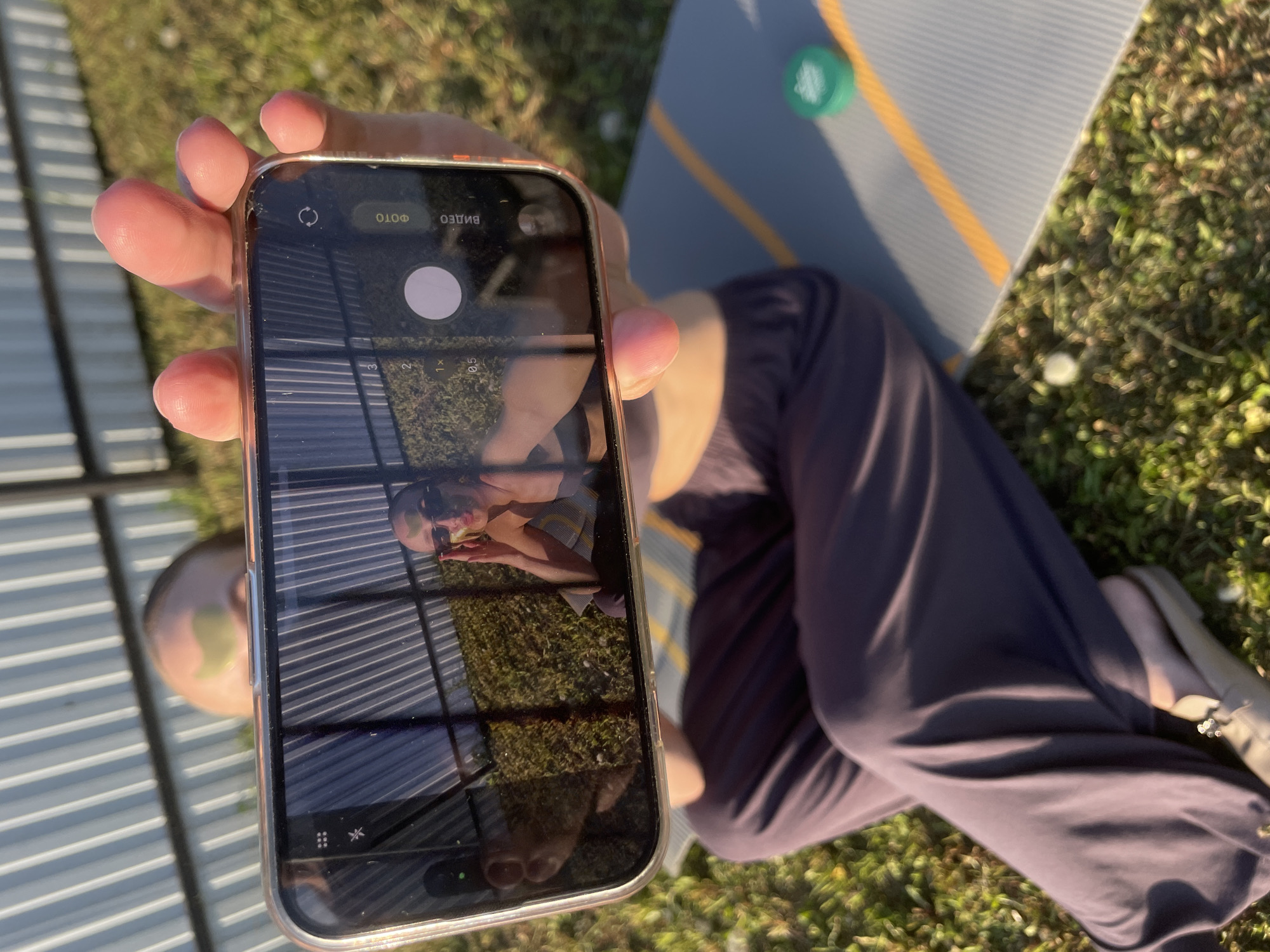 The image shows a person holding a smartphone, displaying its camera app, in front of them. Reflected in the phone screen is an inverted image of the person holding the phone, along with a metal corrugated surface. The person is wearing dark-colored pants. To the right of the phone and person is a yoga mat on grass, with a green object resting on it. The location is Подшивалово, Russia based on the location information.