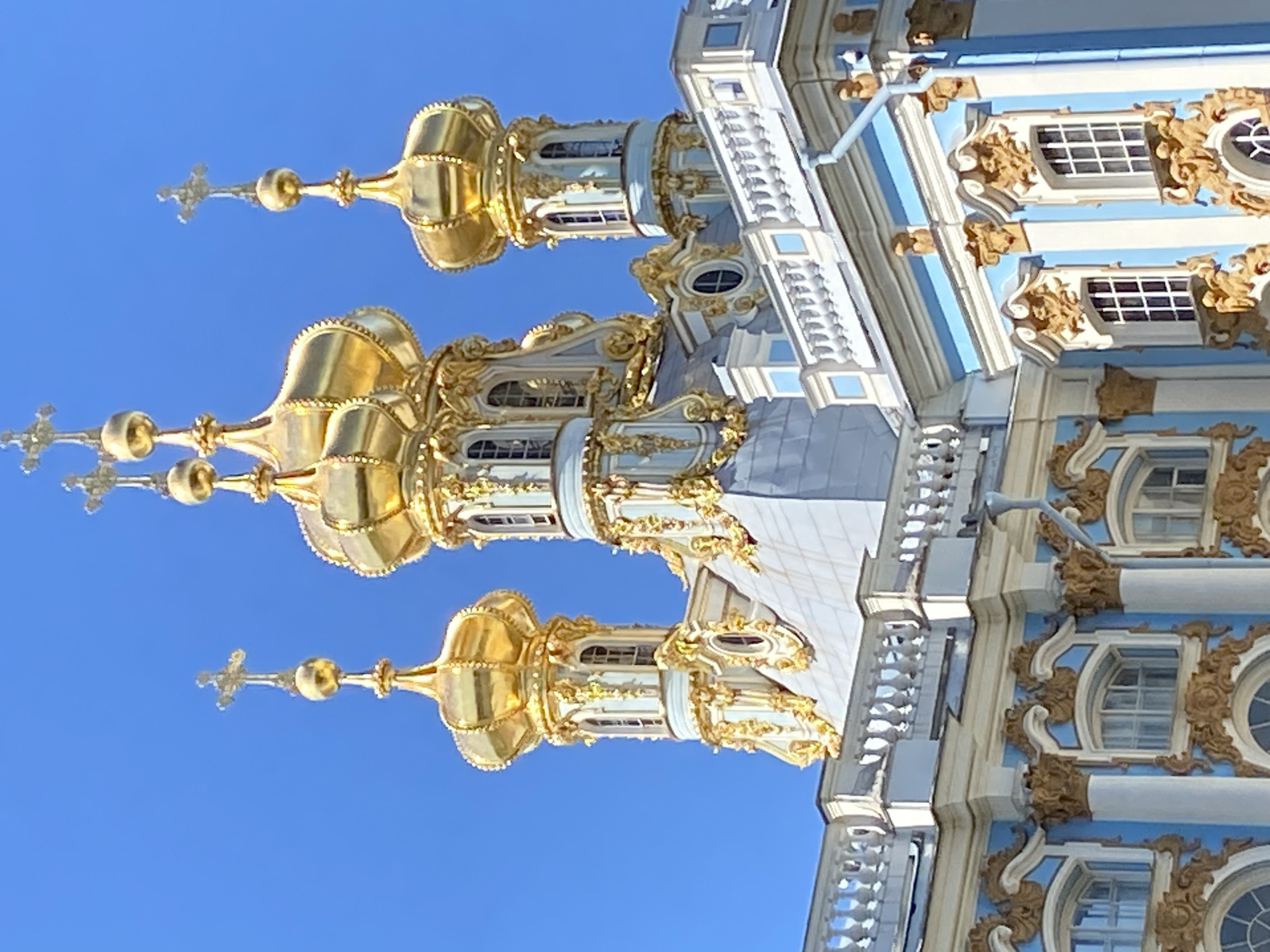 The image shows the Catherine Palace in Pushkin, Russia, under a clear blue sky. The focus is on the ornate golden domes and spires of the palace's architecture, which are topped with crosses. The domes are bulbous and highly decorated with gold ornamentation. Below the domes, the palace facade is visible, featuring a light blue color, white trim, and intricate gold decorative elements around the windows. The windows themselves are rectangular with multiple panes. A white balustrade runs along the upper edge of the visible section of the building. The overall impression is one of grandeur and elaborate baroque or rococo style architecture.