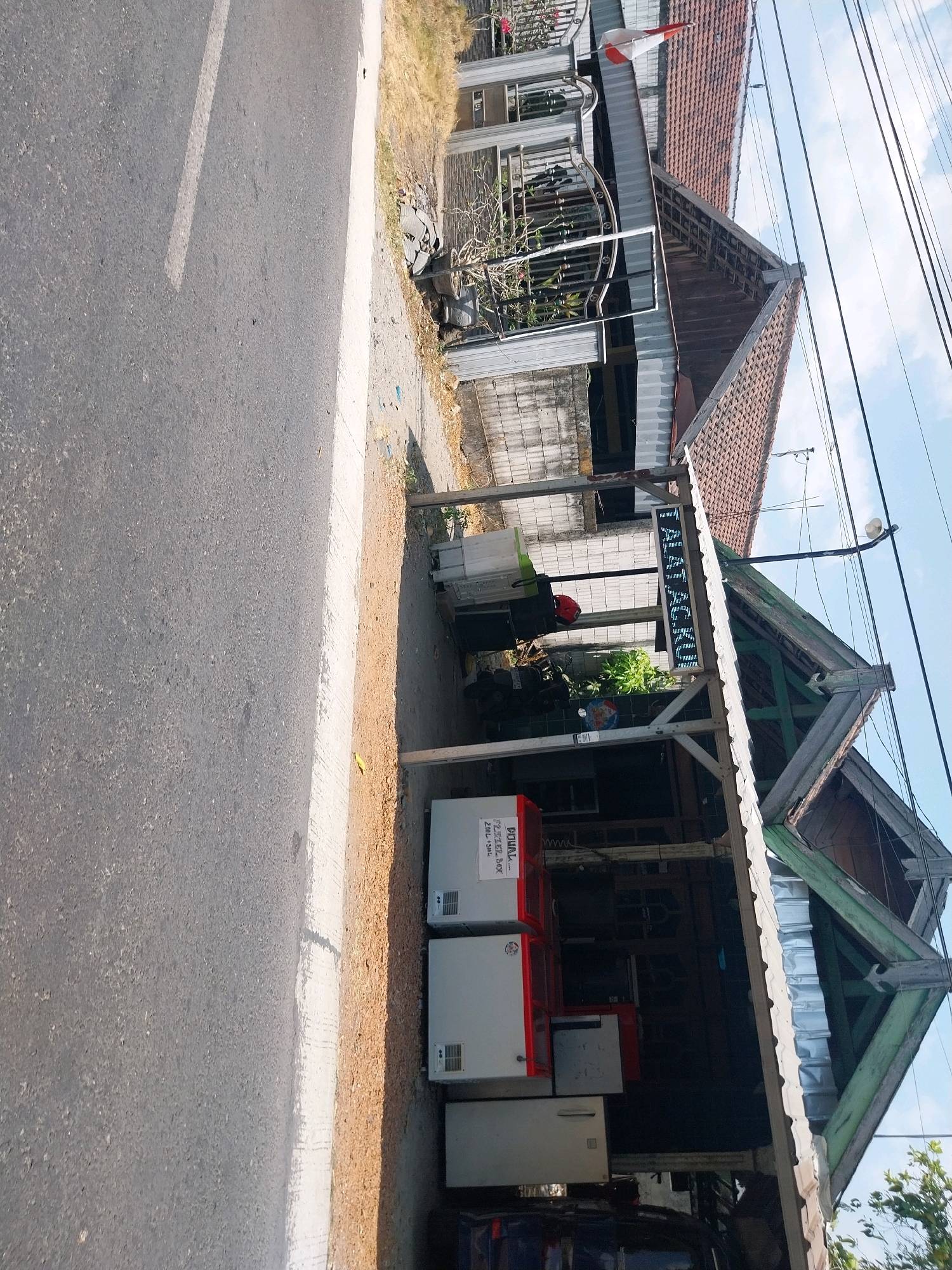 In Campur, Indonesia, a roadside scene unfolds with a focus on commercial activity. The image captures a street with an asphalt surface and a white painted line. Adjacent to the road is a patch of dry grass and dirt. Further back, a building with a red tile roof and supporting pillars is visible. This building appears to be a shop or stall, with a covered area extending outwards. Several appliances, likely refrigerators, are displayed under this covering. One appliance has a red top and a white body, with a label indicating "DIJUAL... F2 FREEZER BOX". Another appliance is entirely white. Above the covered area, a digital sign displays the text "ALAT AC.KU". Electrical wires run overhead against a bright blue sky with some clouds. In the background, a gate and part of another building with a similar tiled roof are present. The overall impression is one of a local business operating in an open-air environment.